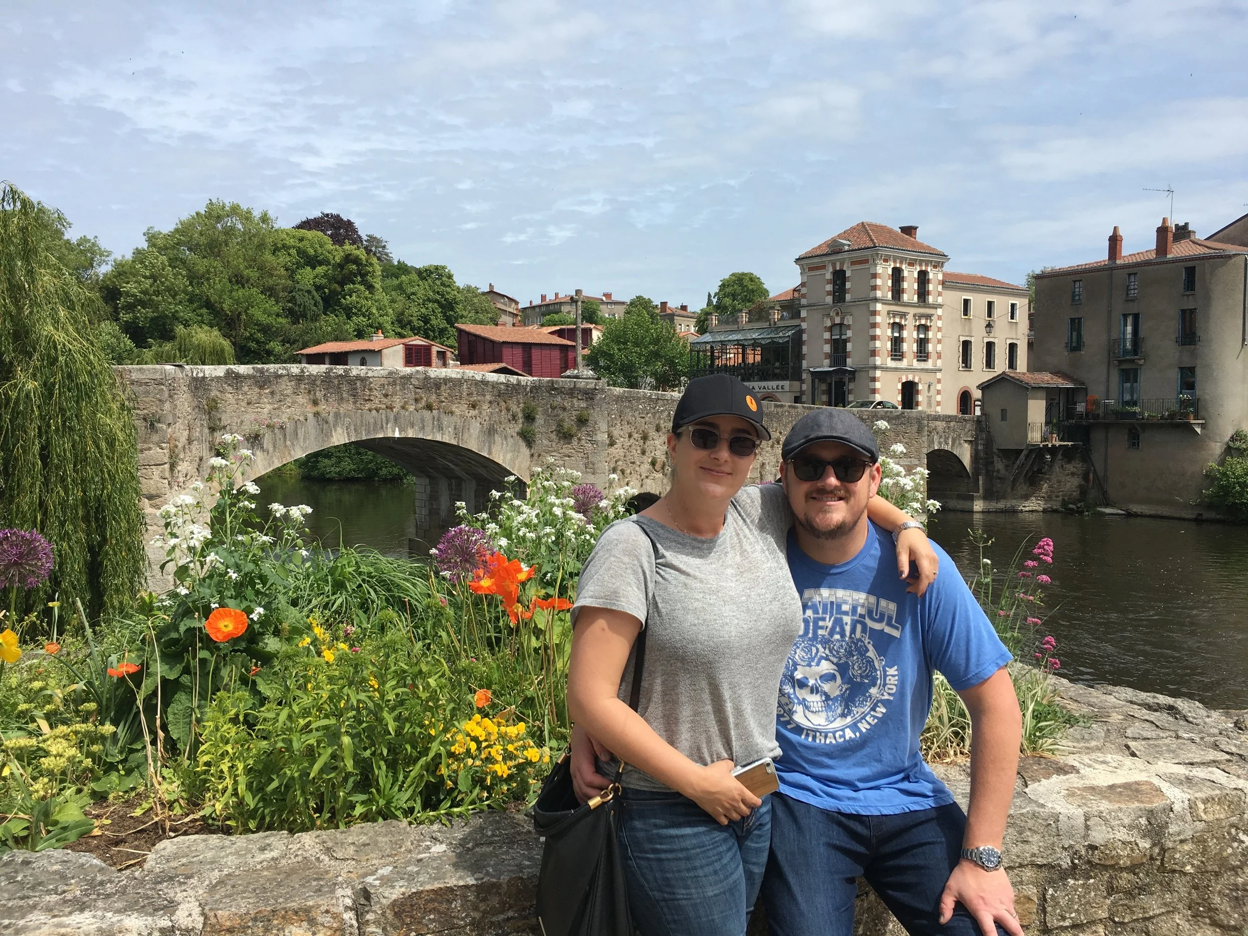 A smiling woman and man pose together outdoors near a stone wall, with a stone bridge, river, colorful flowers, and European-style buildings in the background.