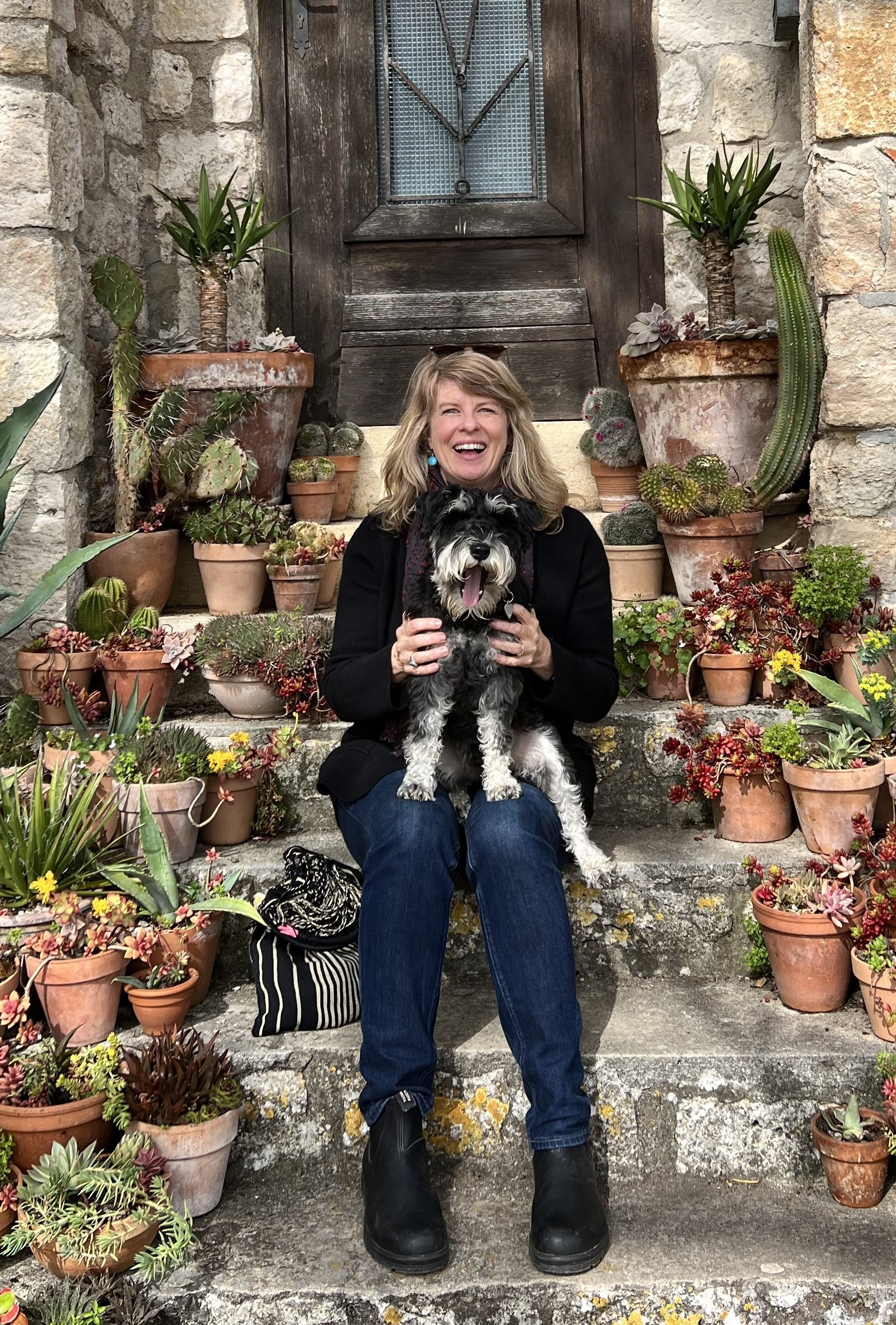 A woman sitting on outdoor stone steps holding a black and white dog, surrounded by potted succulents and cacti, with a rustic wooden door in the background.