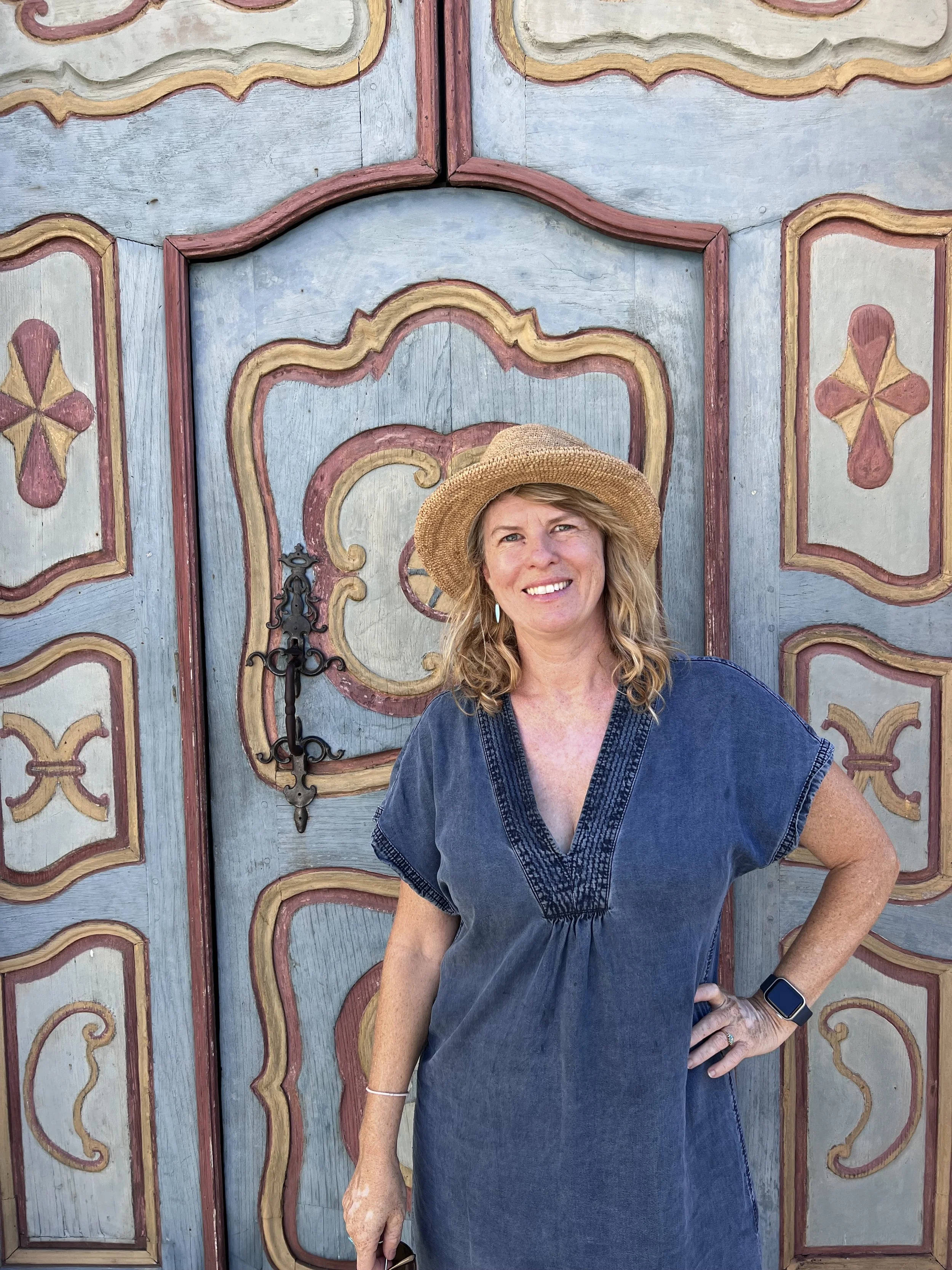 A woman with wavy blonde hair wearing a straw hat, denim dress, and smartwatch standing in front of an ornate painted wooden door.