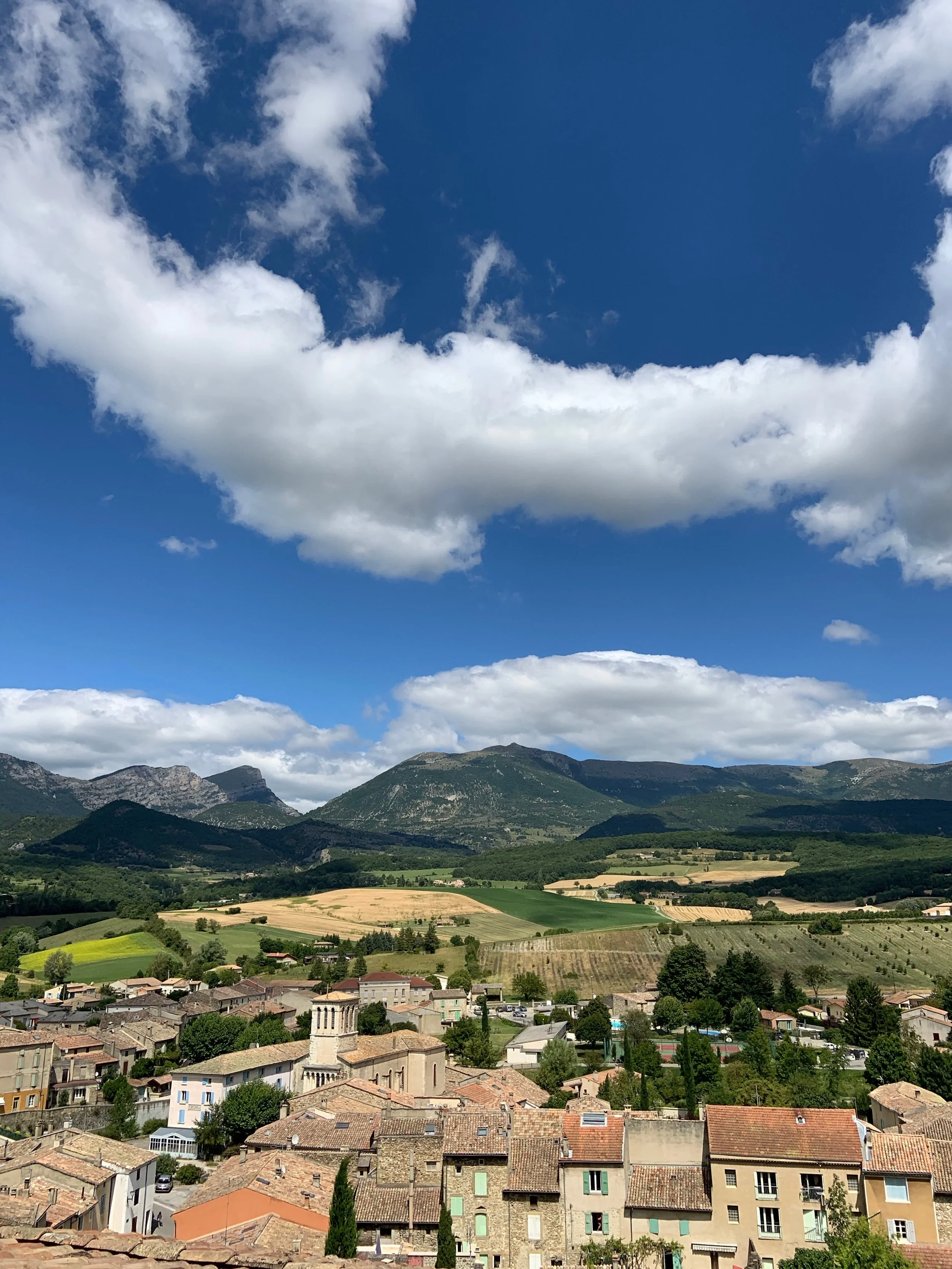 A panoramic view of a small village with old buildings and red-tiled roofs, set against rolling green hills and mountains under a partly cloudy sky.