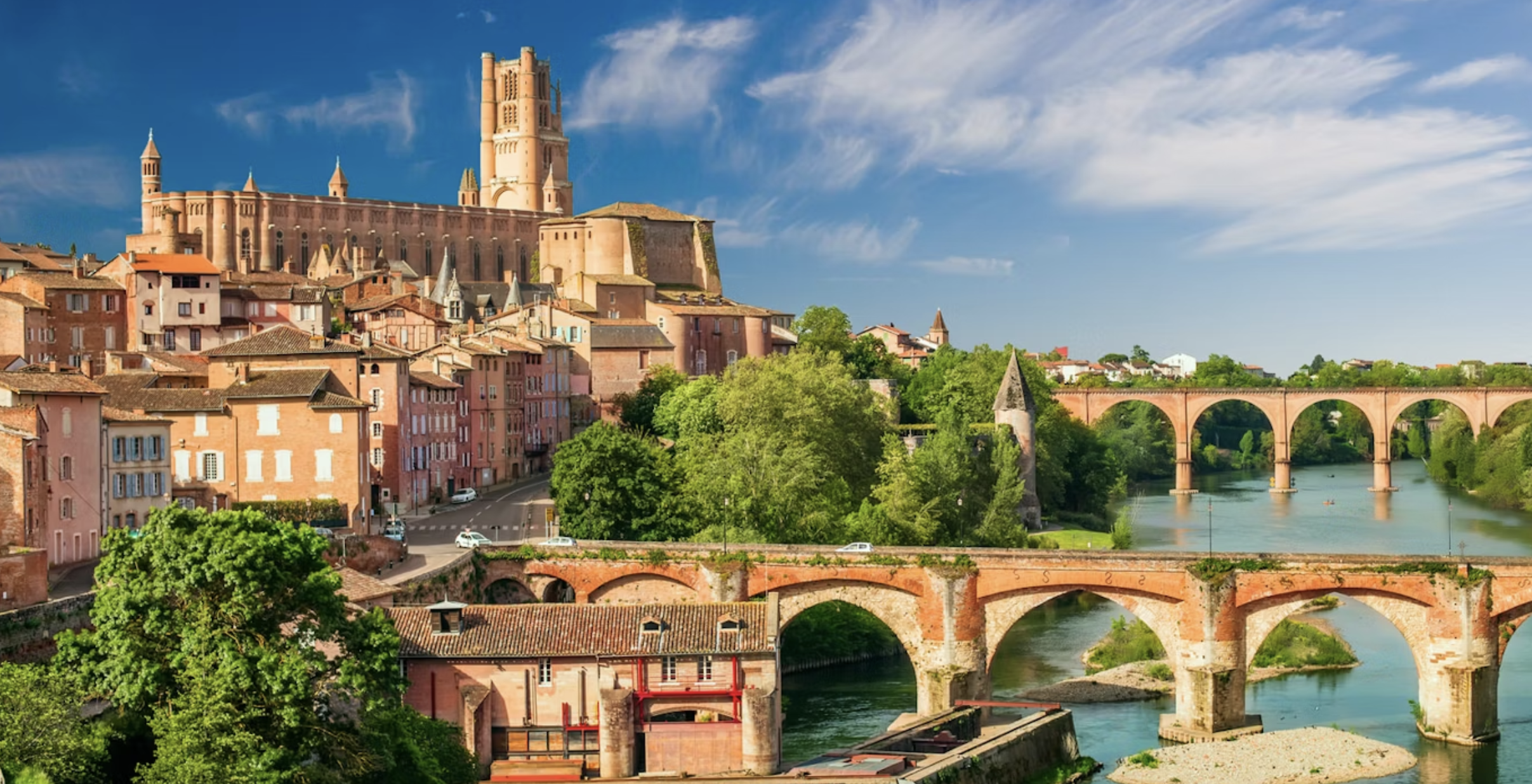 Albi - Scenic view of a historic European town on a hill, featuring a large castle at the top, surrounded by colorful buildings, lush green trees, and a river with multiple brick and stone bridges crossing over it, under a partly cloudy blue sky.