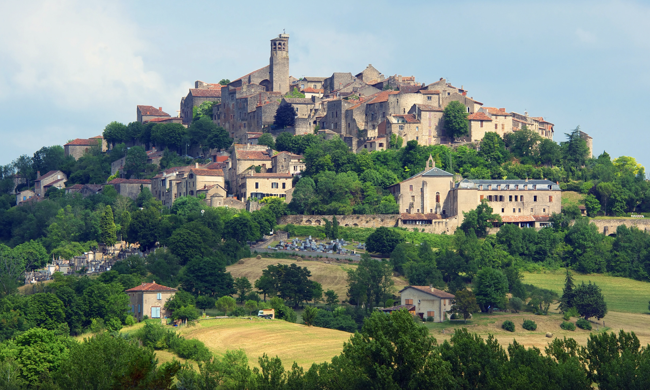Cordes-sur-Ciel - a historic hilltop village with stone buildings and a tower, surrounded by green trees and rolling fields.