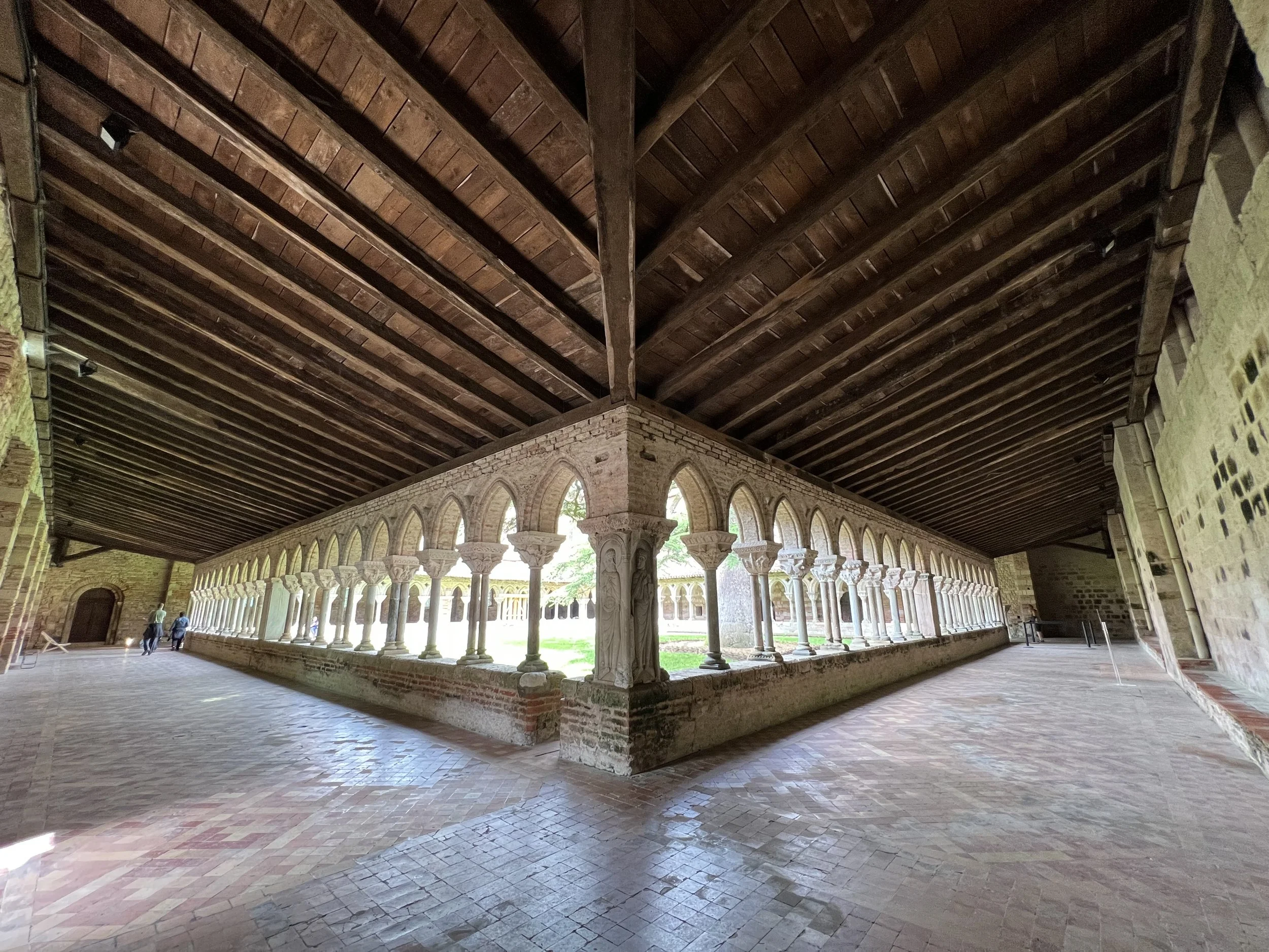 Tour historical abbeys. Interior view of a historic cloister with a wooden ceiling, brick walls, and a stone walkway. The cloister features a series of arches supported by columns, with a view of the courtyard outside visible through the arches.