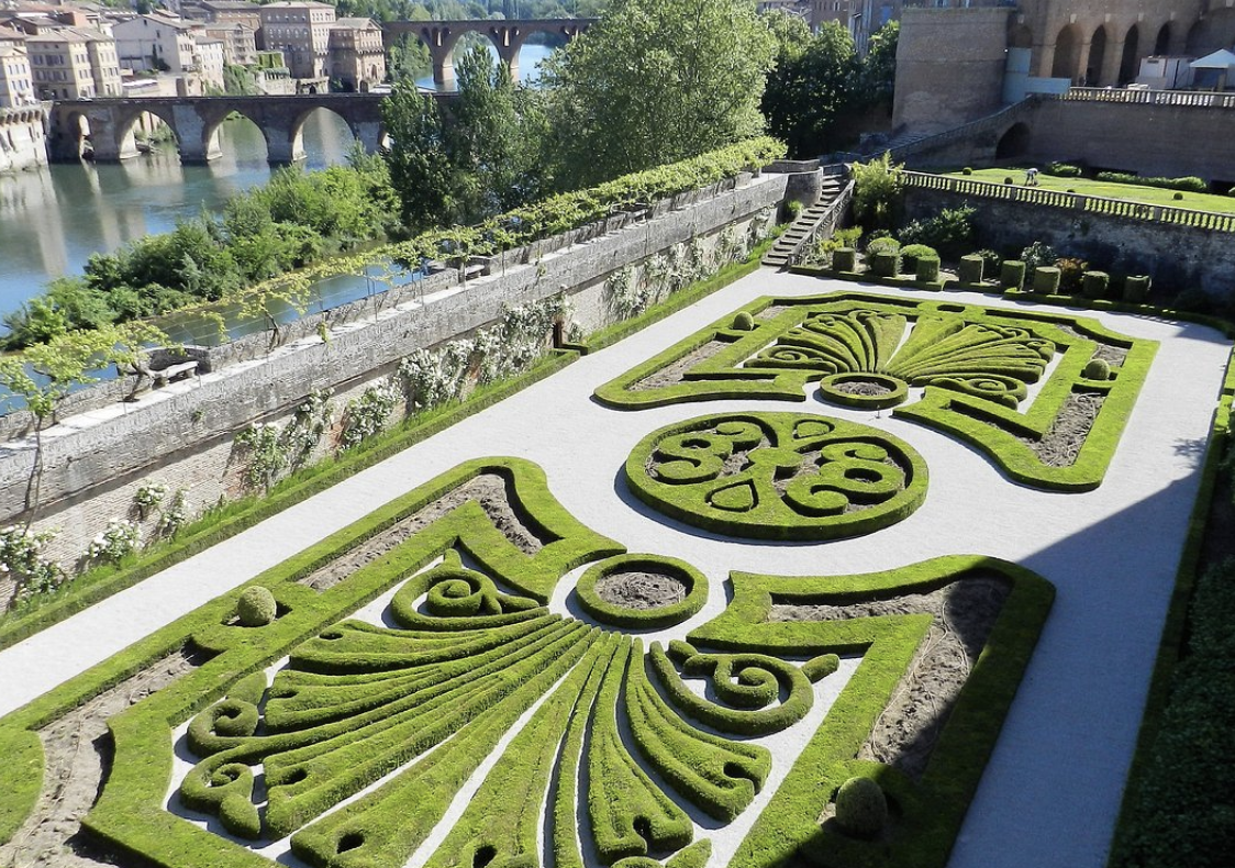 Albi discoveries - A beautifully manicured garden with intricate, symmetrical green topiary design, bordered by white gravel, situated near a river with stone bridges and historic buildings in the background.