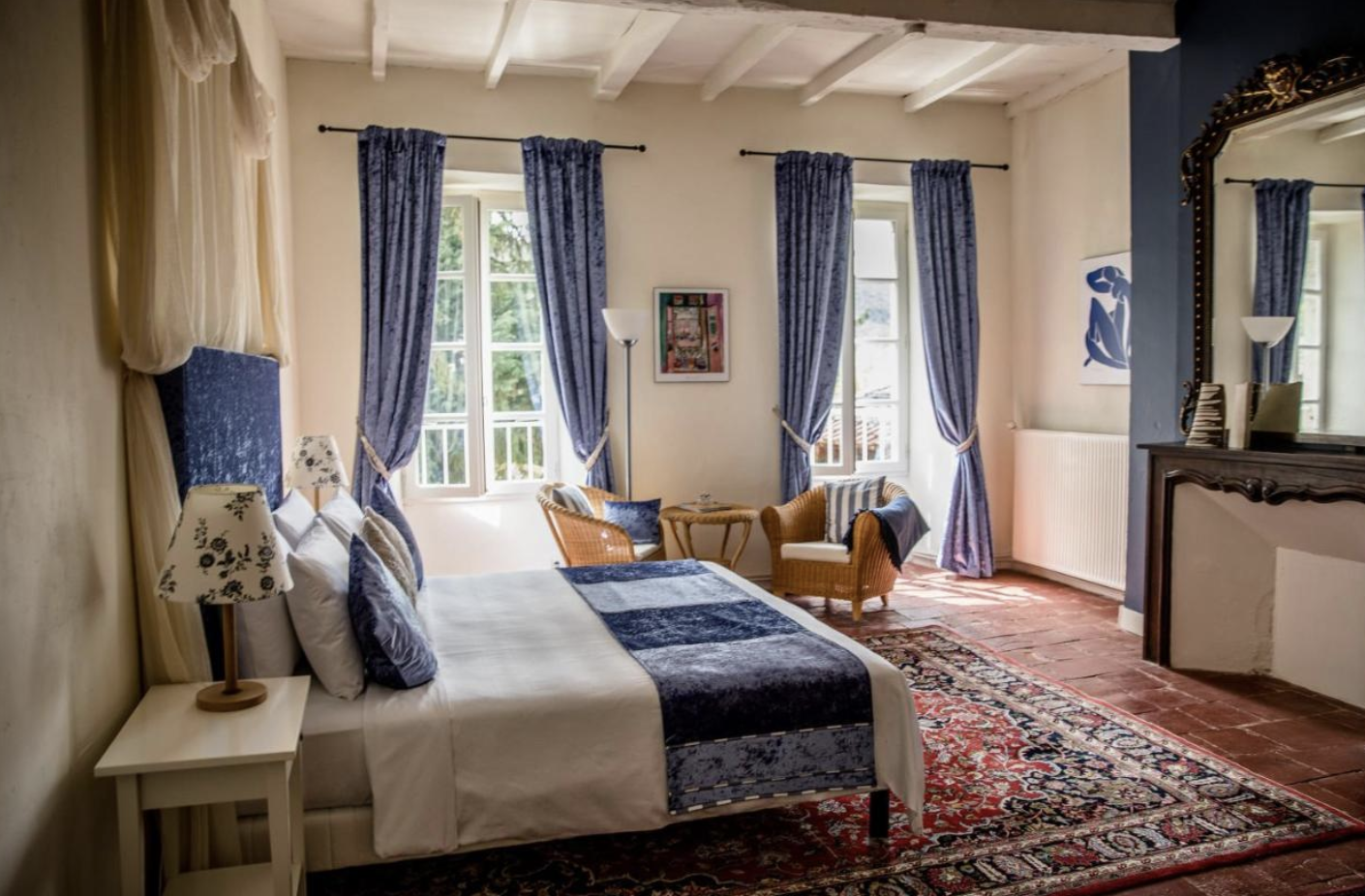 Bright bedroom with large windows, blue curtains, a white bed with pillows and a blue bed runner, a white bedside table with a patterned lamp, wicker chairs, a floral rug, a floor lamp, and a large ornate mirror.