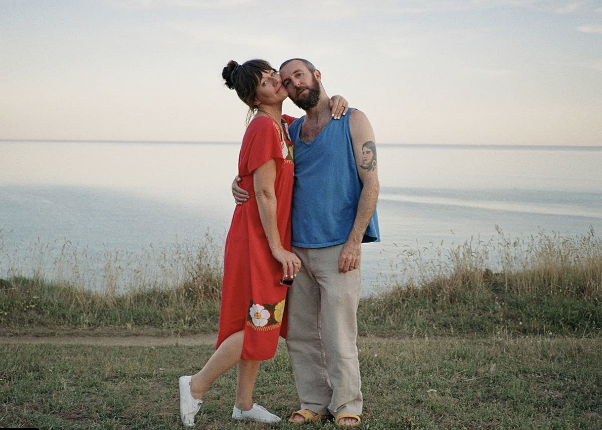 A woman in a red dress and a man in a blue tank top and beige pants cuddling outdoors near a body of water during sunset.