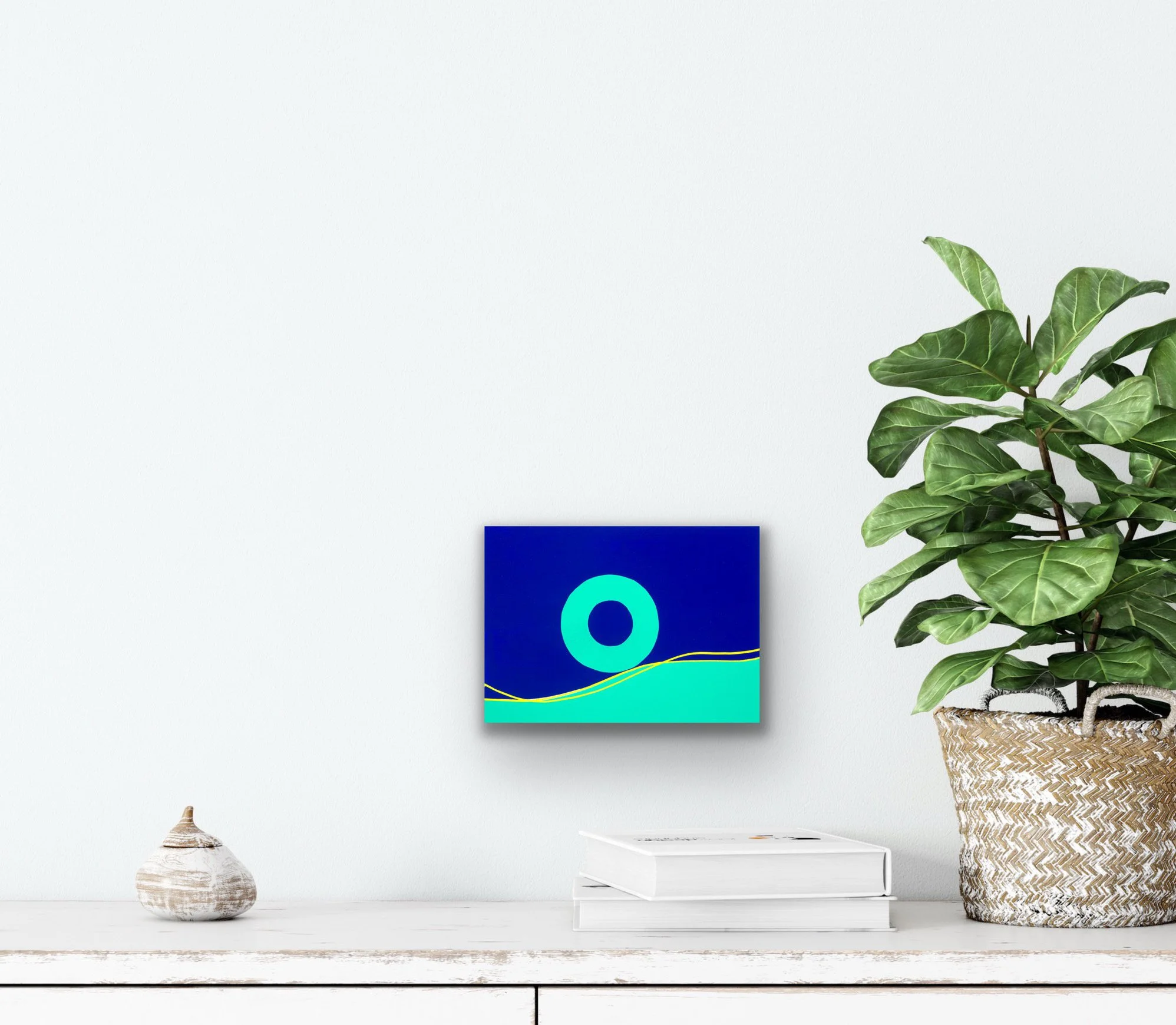 “Gravitation by Paula Cahill displayed in a minimal interior with a plant and books.”G