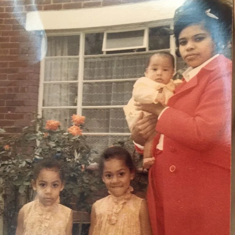 A woman in a red suit holding a baby, with two young girls standing in front of her, in front of a window with a brick wall and orange flowers in the background.