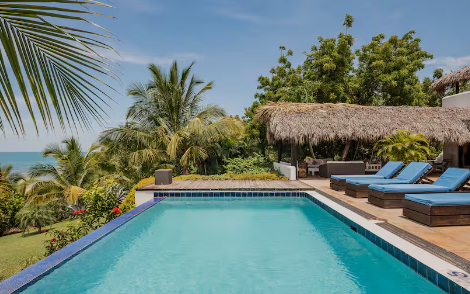 Swimming pool at a tropical resort with lounge chairs and a thatched-roof cabana, surrounded by lush greenery and palm trees