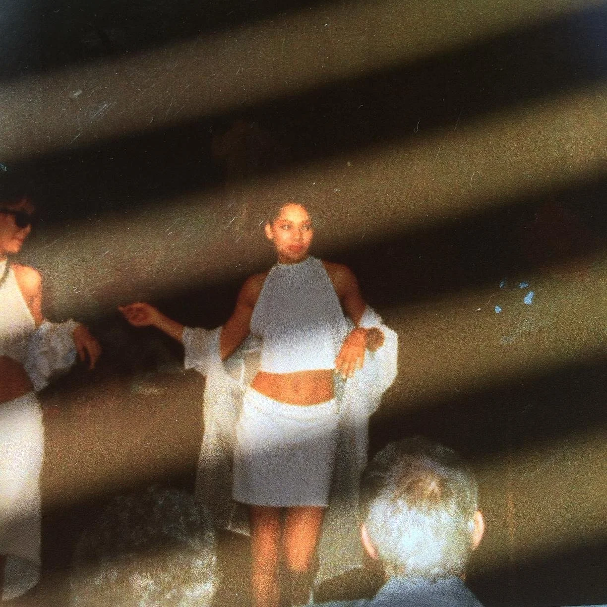 A woman with dark hair wearing a white high-neck top and a white skirt, holding a white cloth, standing in a dark indoor space with audience members observing.