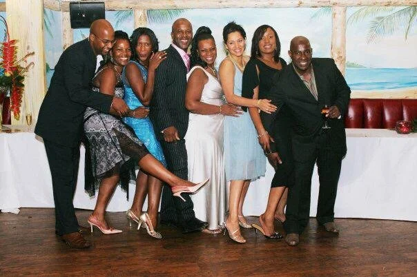 Group of nine people dressed in formal attire at a celebration event, standing in front of a tropical-themed backdrop.