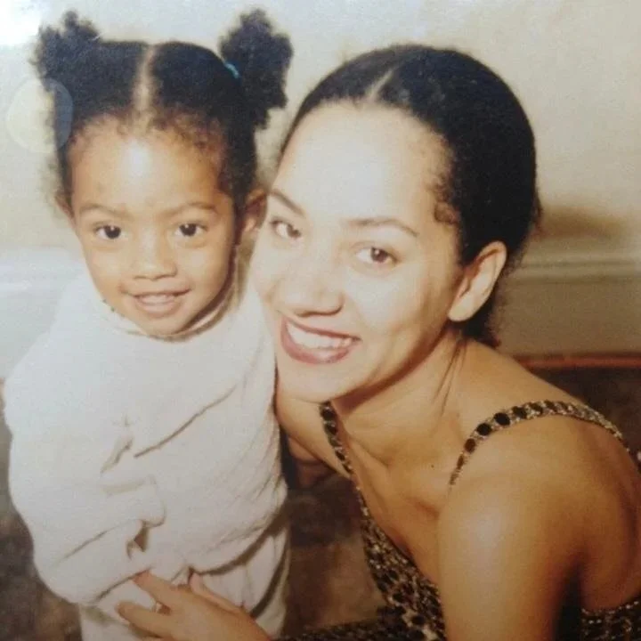 A woman and a young girl smiling and posing for a photo together, with the woman wearing a leopard print dress and the girl wearing a light-colored top, indoors against a neutral wall.