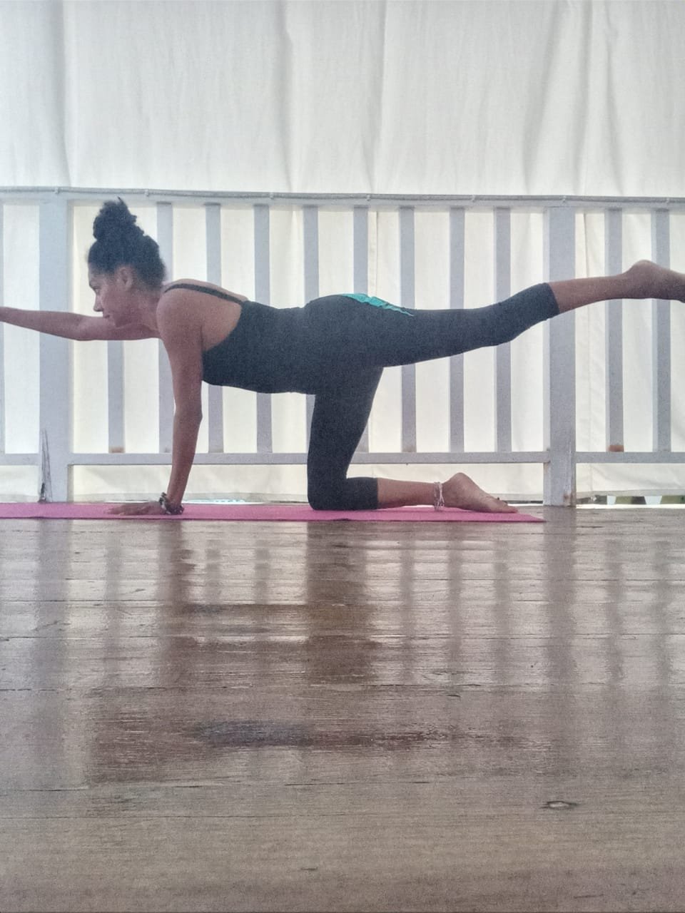 A woman practicing yoga on a pink mat, balancing on her left hand and right knee with her left arm and right leg extended outward.