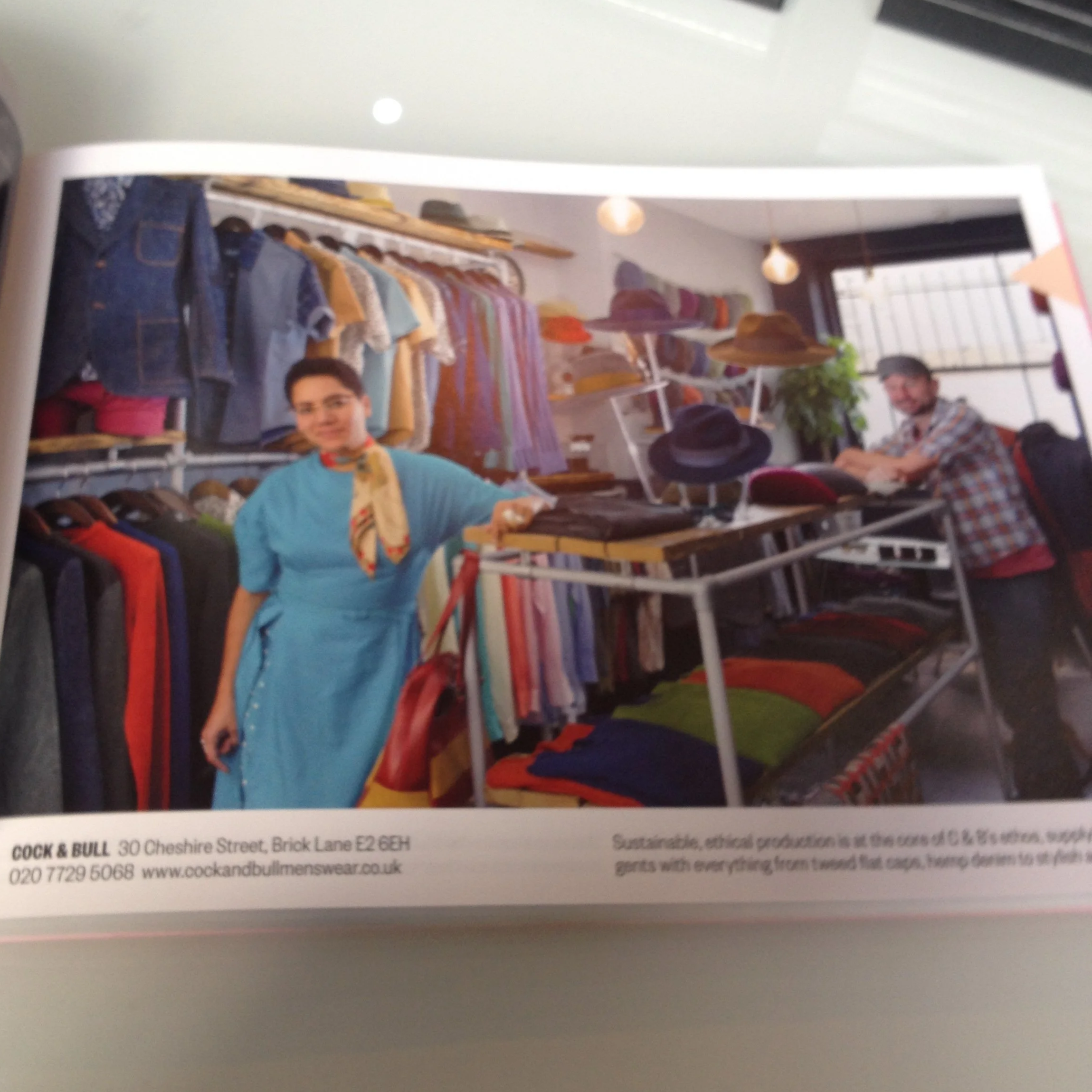 Photo of a clothing store with two people; a woman in a blue dress standing near clothing racks and a man seated at a table with hats and accessories, with shelves of folded clothes and hats in the background.