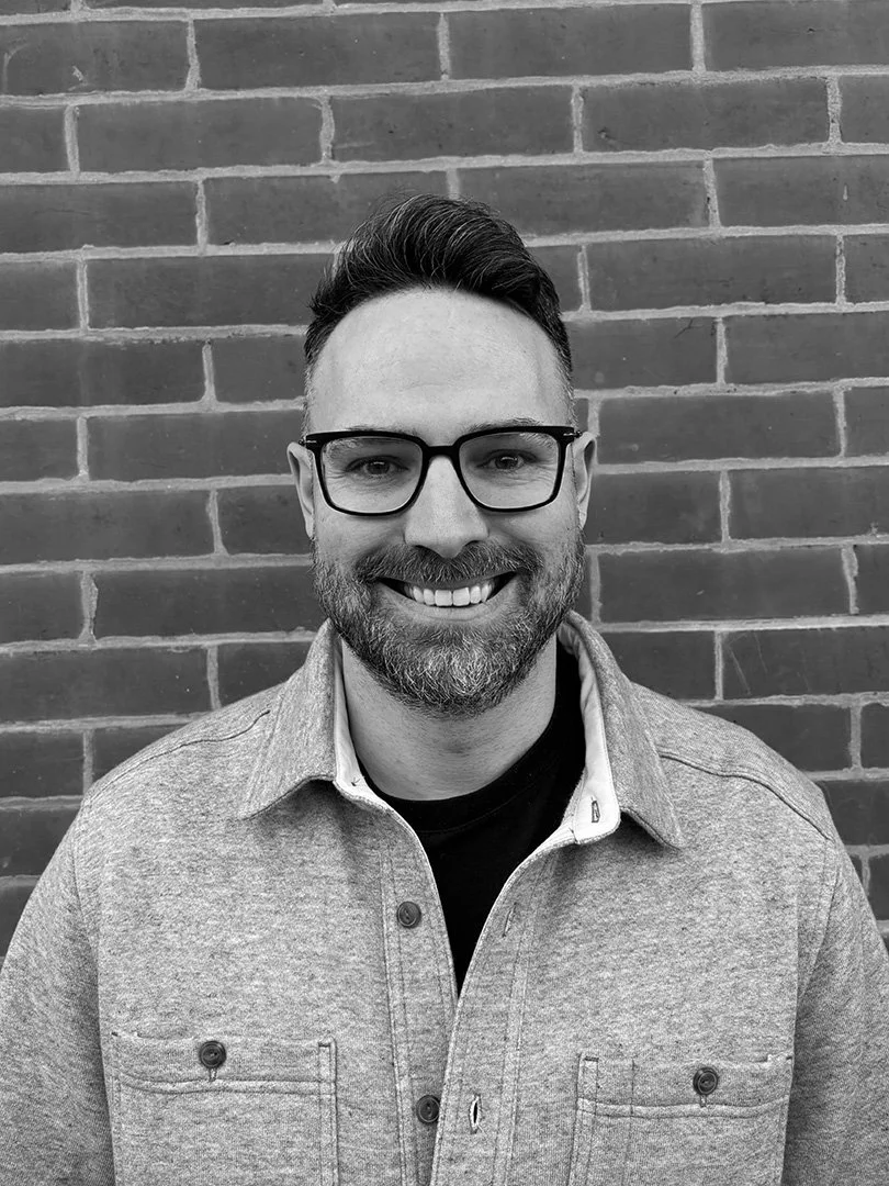 A smiling man with glasses, a beard, and styled hair, wearing a gray button-up shirt layered over a black shirt, standing in front of a brick wall.