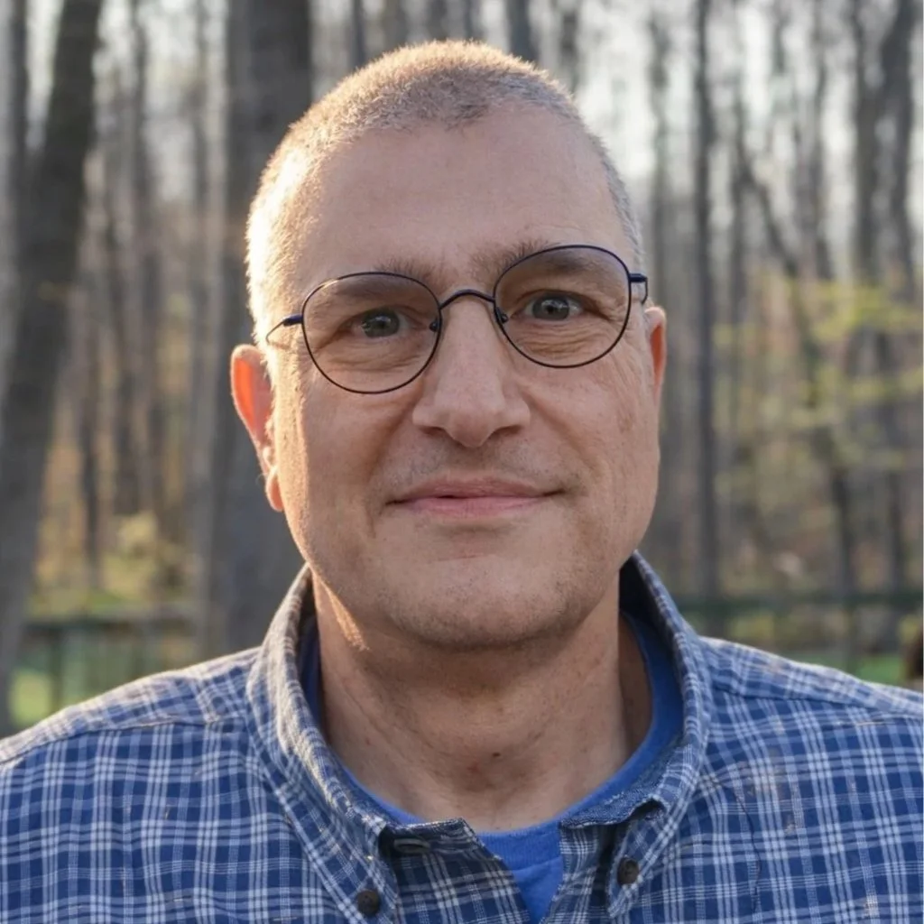 A man wearing glasses and a plaid shirt outdoors in a wooded area.