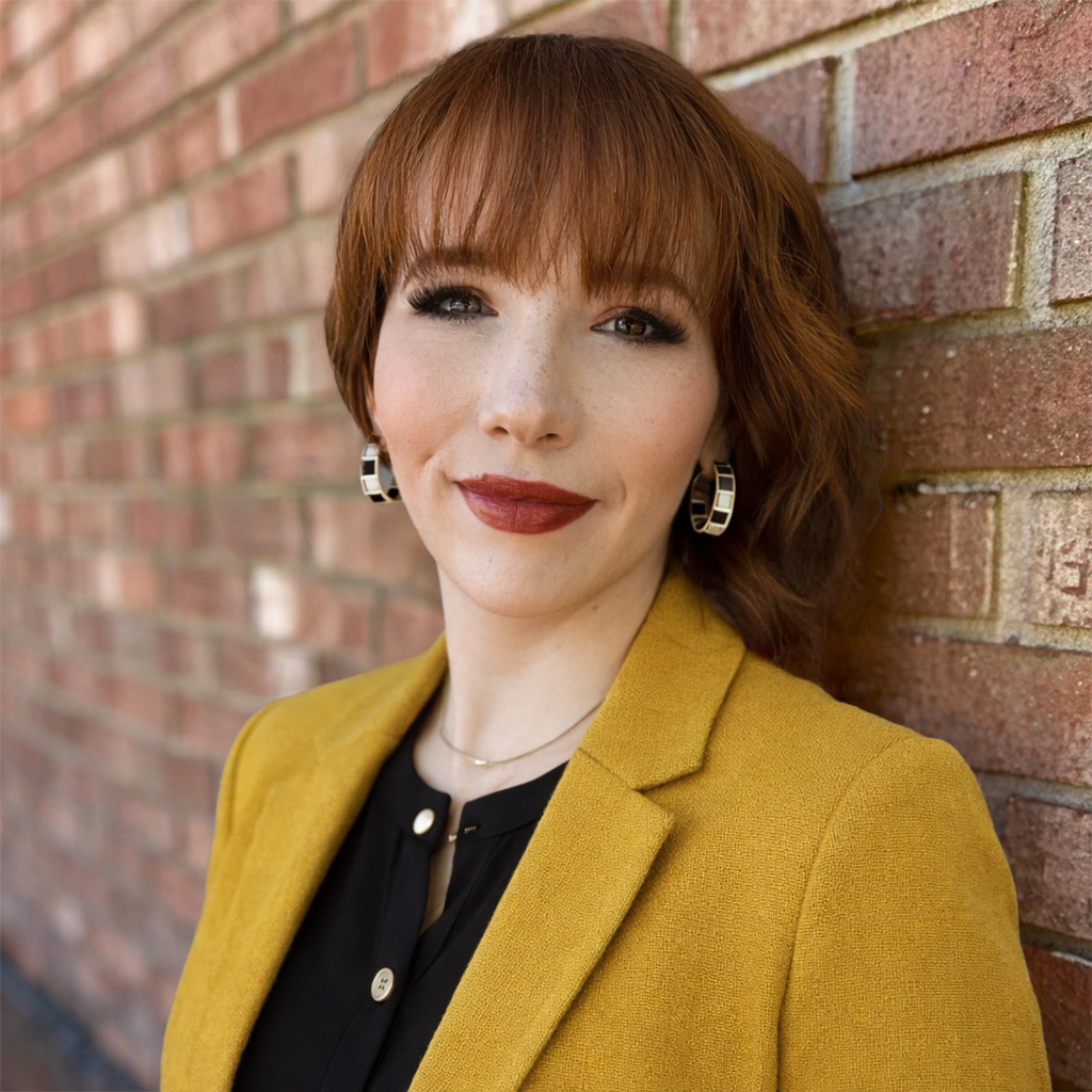 A woman with red hair and fair skin leaning against a brick wall, wearing a yellow blazer and black top, with earrings and red lipstick, outdoors.