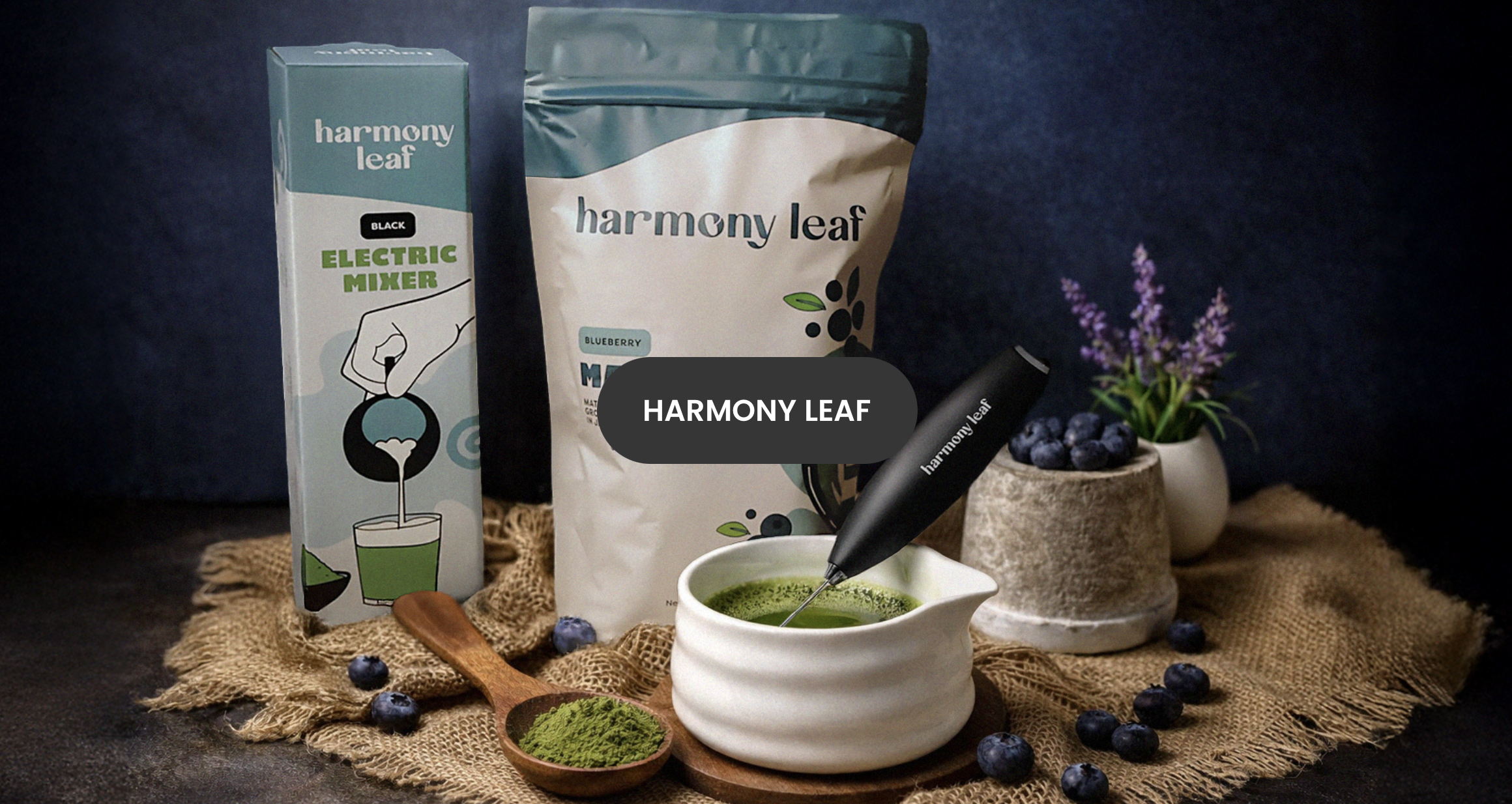 Various Harmony Leaf tea products including a box of electric mixer, a bag of blue berry matcha, a white ceramic bowl with green matcha tea, a black Matcha whisk, a potted plant with purple flowers, and scattered blueberries on a burlap cloth.