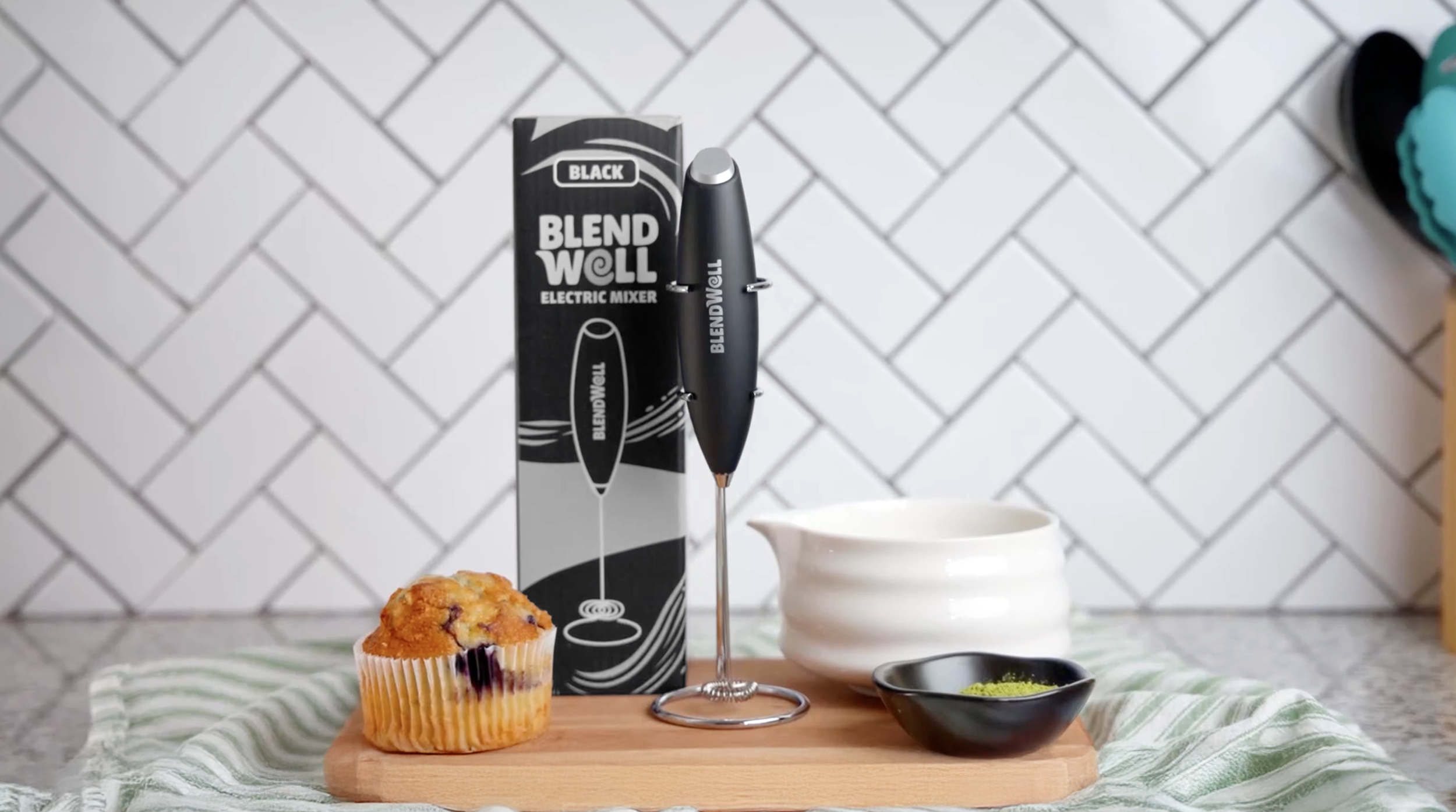 A muffin, a black electric smoothie blender with its box, a white ceramic pitcher, and a small black bowl with green powder are placed on a wooden board on a green and white striped cloth, with a tiled backsplash in the background.