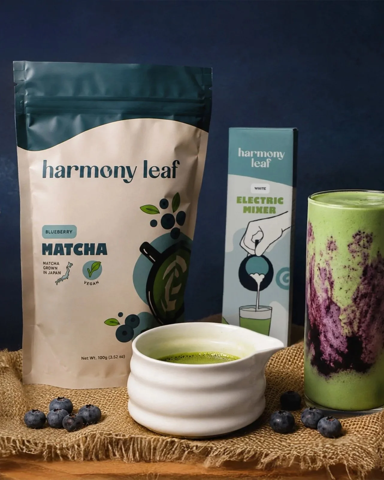 A person is stirring a matcha green tea latte in a white cup with a white spoon labeled 'harmony leaf.' There is a bag of blueberry matcha on the table, with blueberries scattered around, and a blue background with a starry sky pattern.
