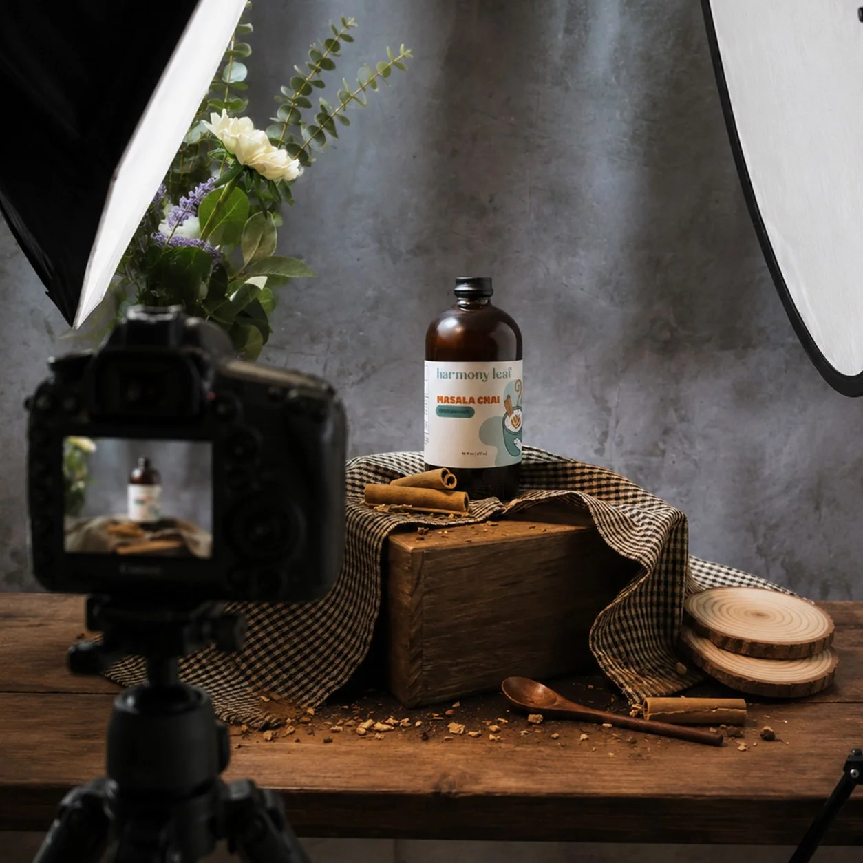 Behind-the-scenes product photography of a Harmony Leaf Masala Chai bottle on a styled wooden set with a camera, studio lights, cinnamon sticks, and floral accents