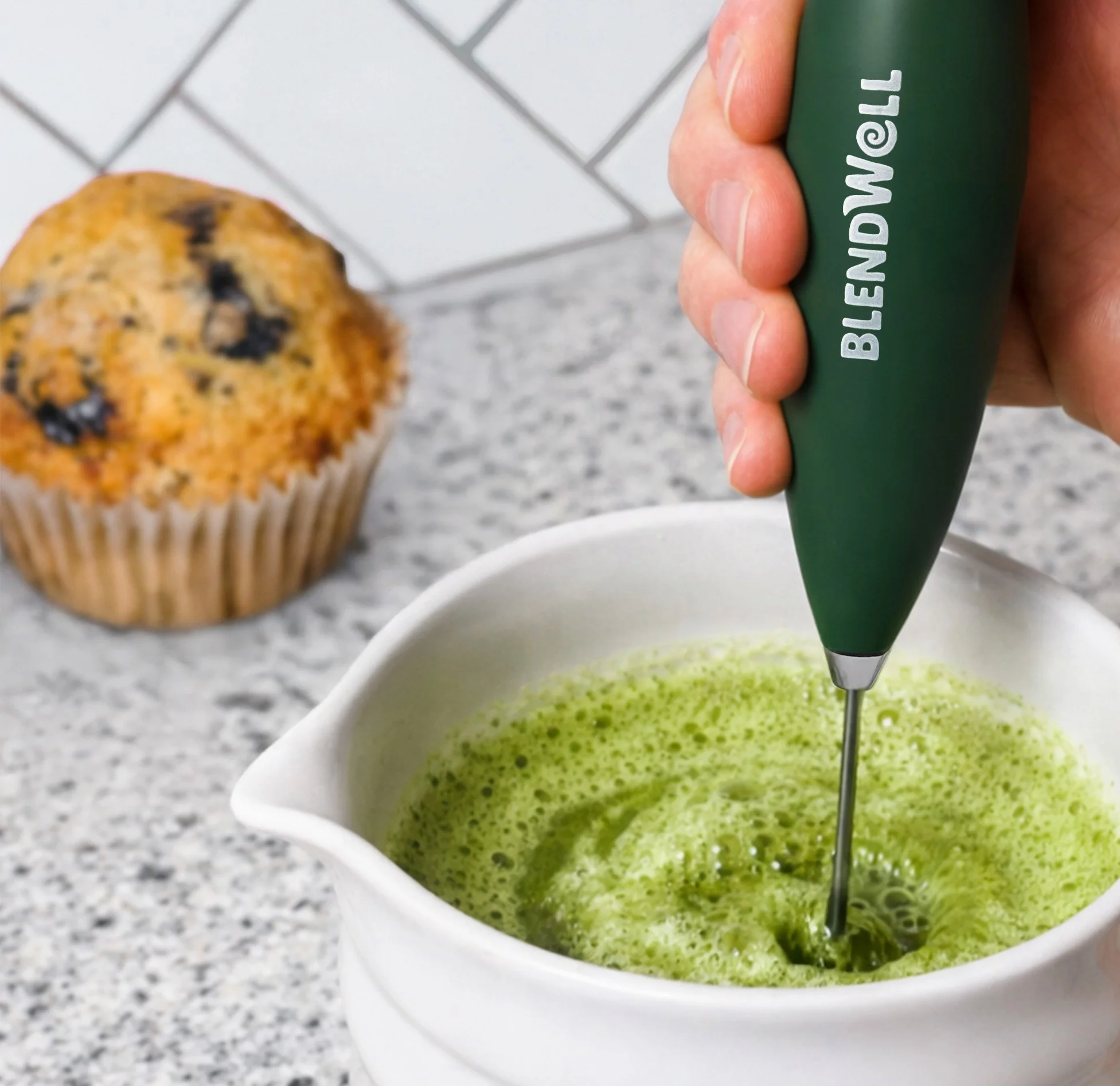 Person using a hand-held blender to blend green liquid in a white bowl, with a blueberry muffin in the background on a speckled gray countertop.