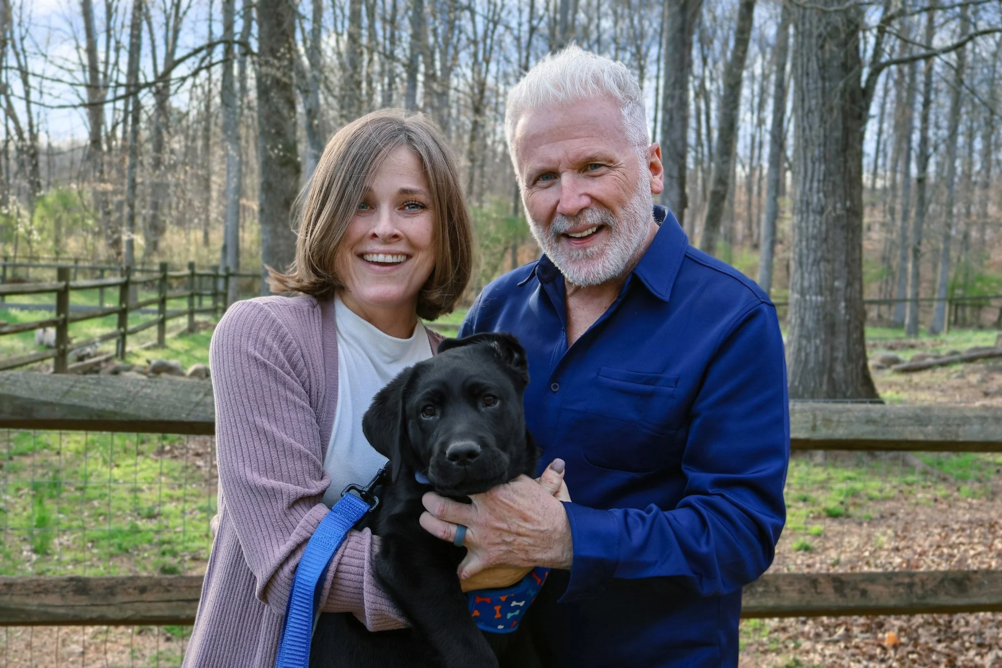 A happy middle-aged man and woman smiling outdoors holding a black puppy in front of a wooden fence and trees.