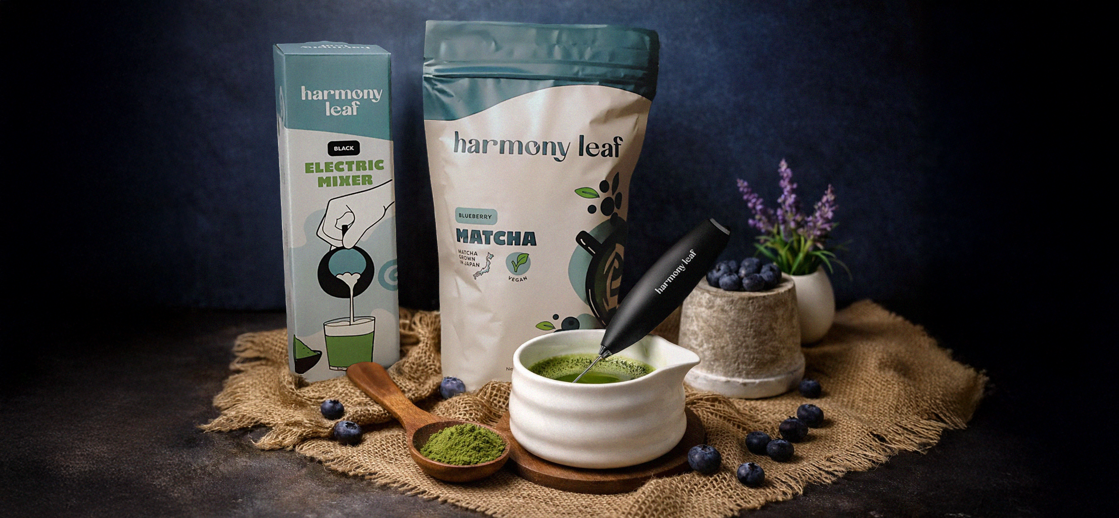 Arrangement of Harmony Leaf matcha products including a packet, a box of electric mixer, a bowl of prepared matcha, a spoon of powdered matcha, and blueberries, with purple flowers in a small white vase on a burlap cloth background.