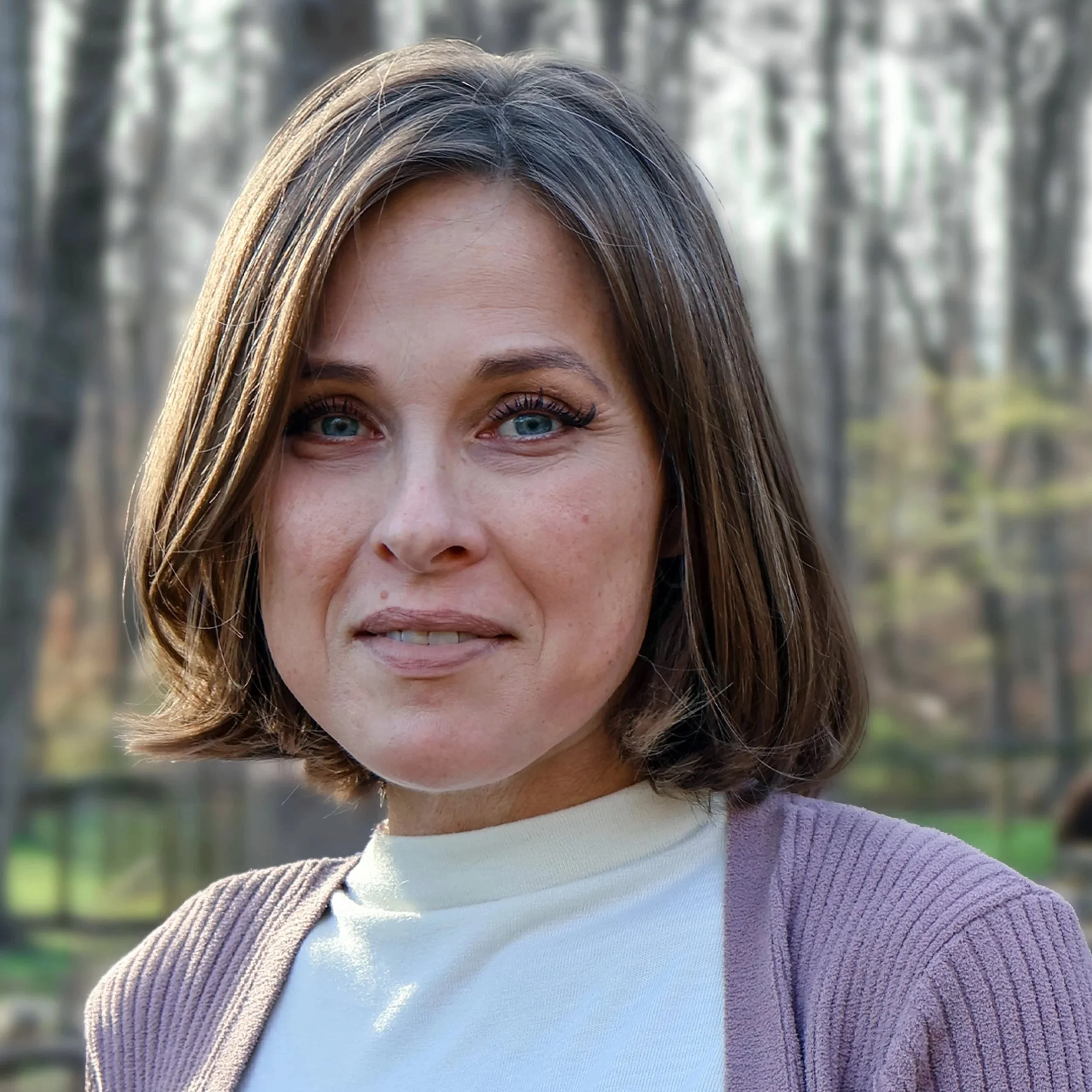 A woman with shoulder-length brown hair and blue eyes standing outdoors with trees in the background.