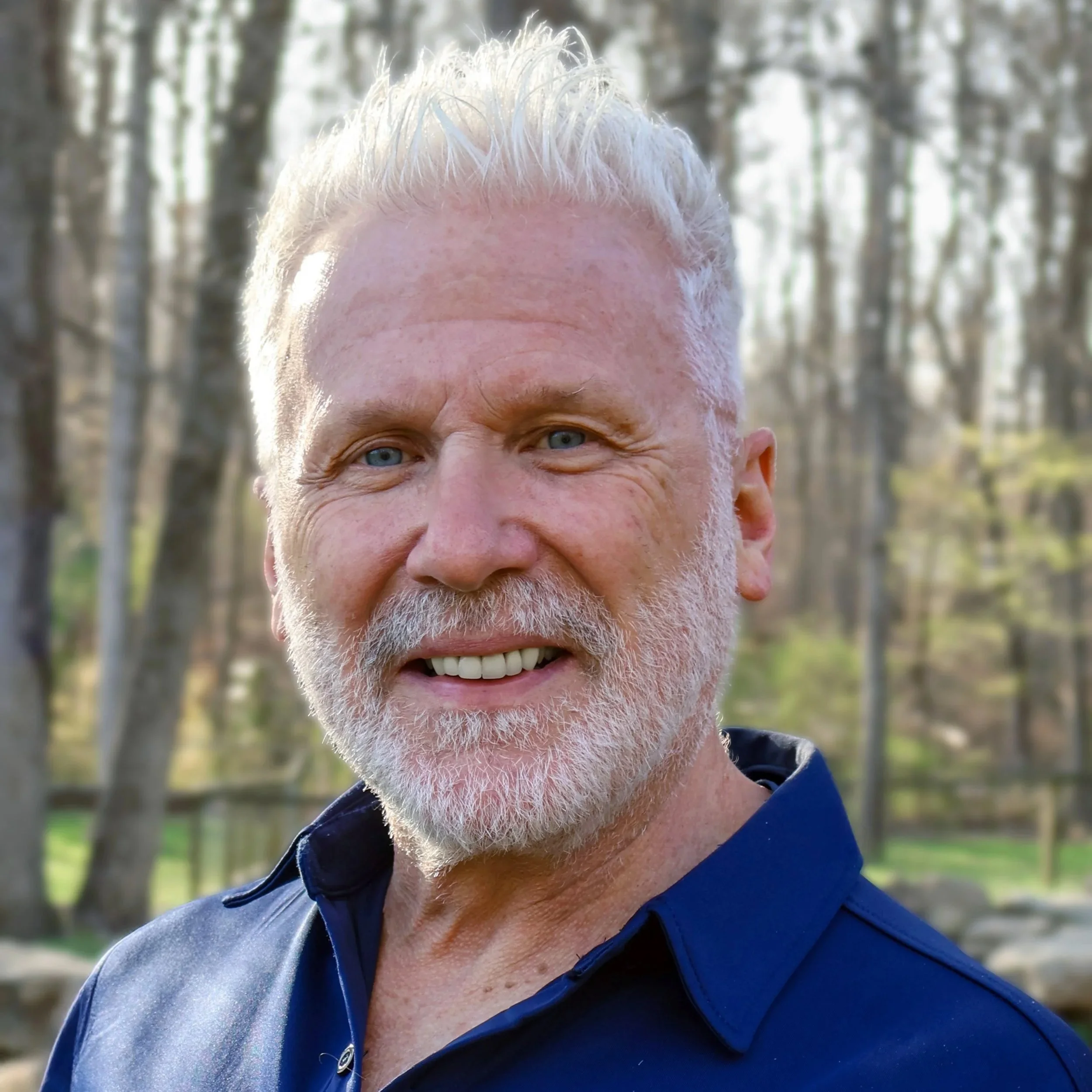 Close-up of a smiling older man with white hair and beard, wearing a navy blue shirt, outdoors with a blurred background of trees and greenery.