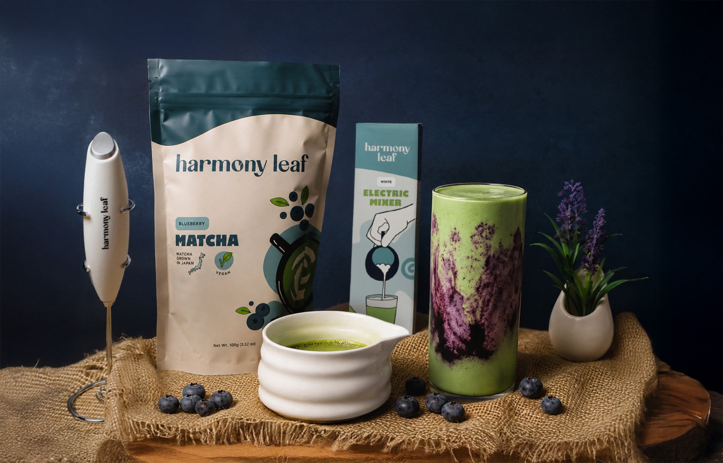 Arrangement of Harmony Leaf matcha products including a packet, a box of electric mixer, a bowl of prepared matcha, a spoon of powdered matcha, and blueberries, with purple flowers in a small white vase on a burlap cloth background.