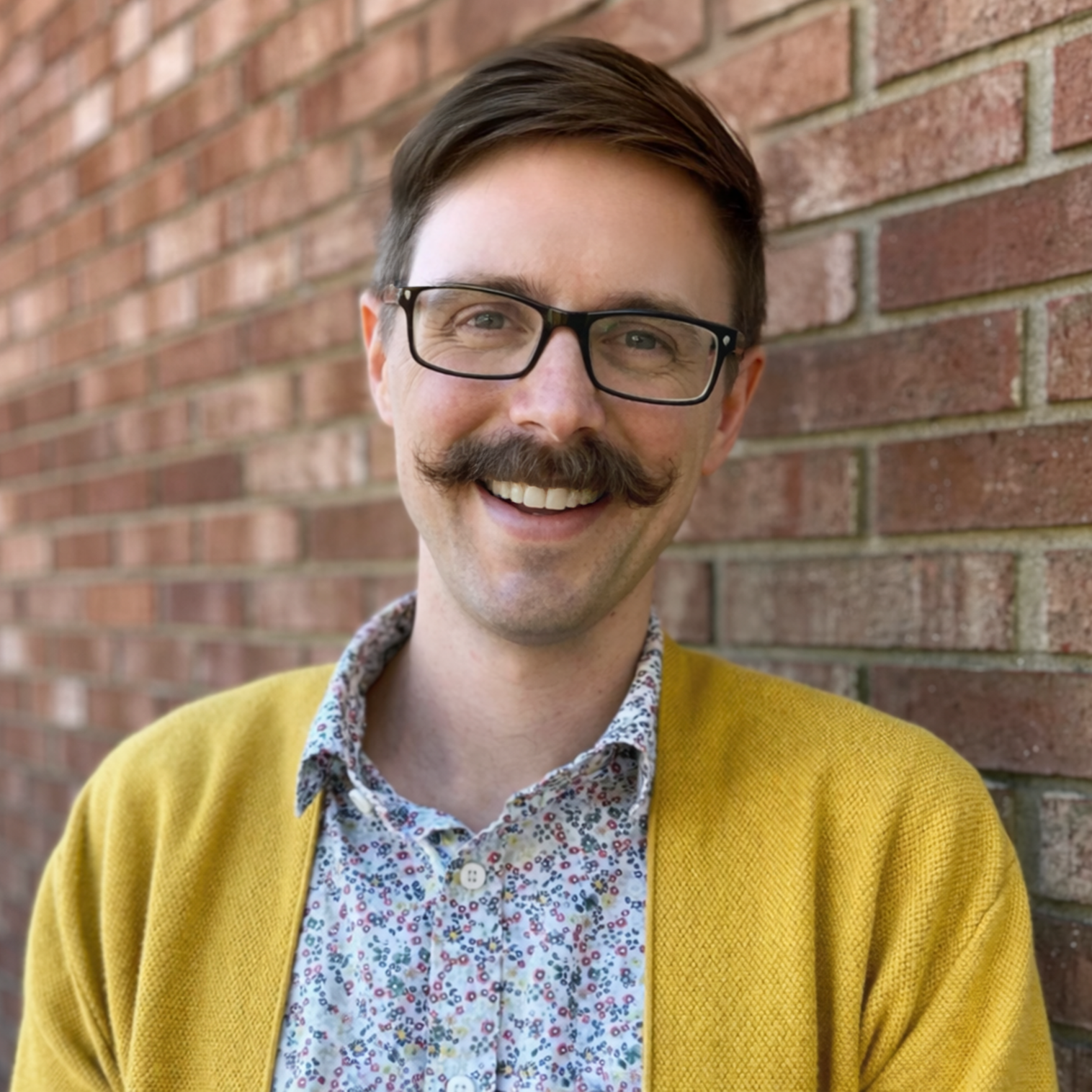 A man with glasses and a mustache smiling, standing against a brick wall.