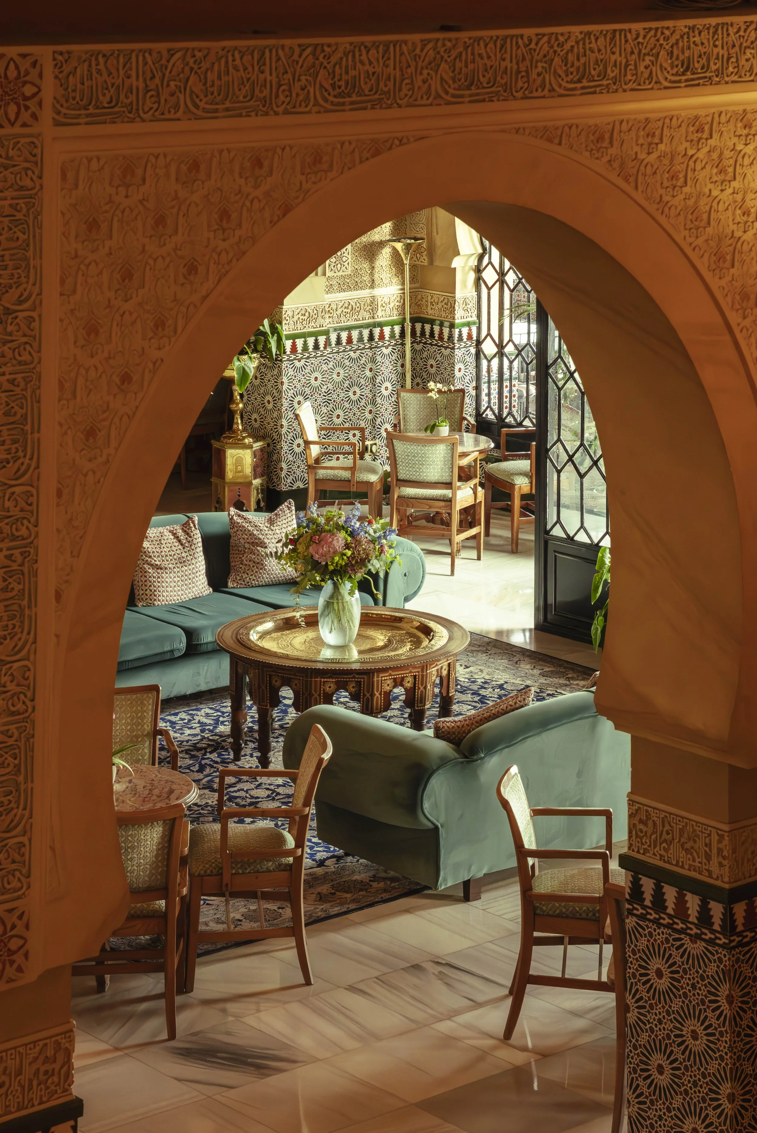 Salón decorado con muebles elegantes, cojines, un florero con flores en una mesa y paredes con detalles árabes, vista a través de un arco ornamentado.