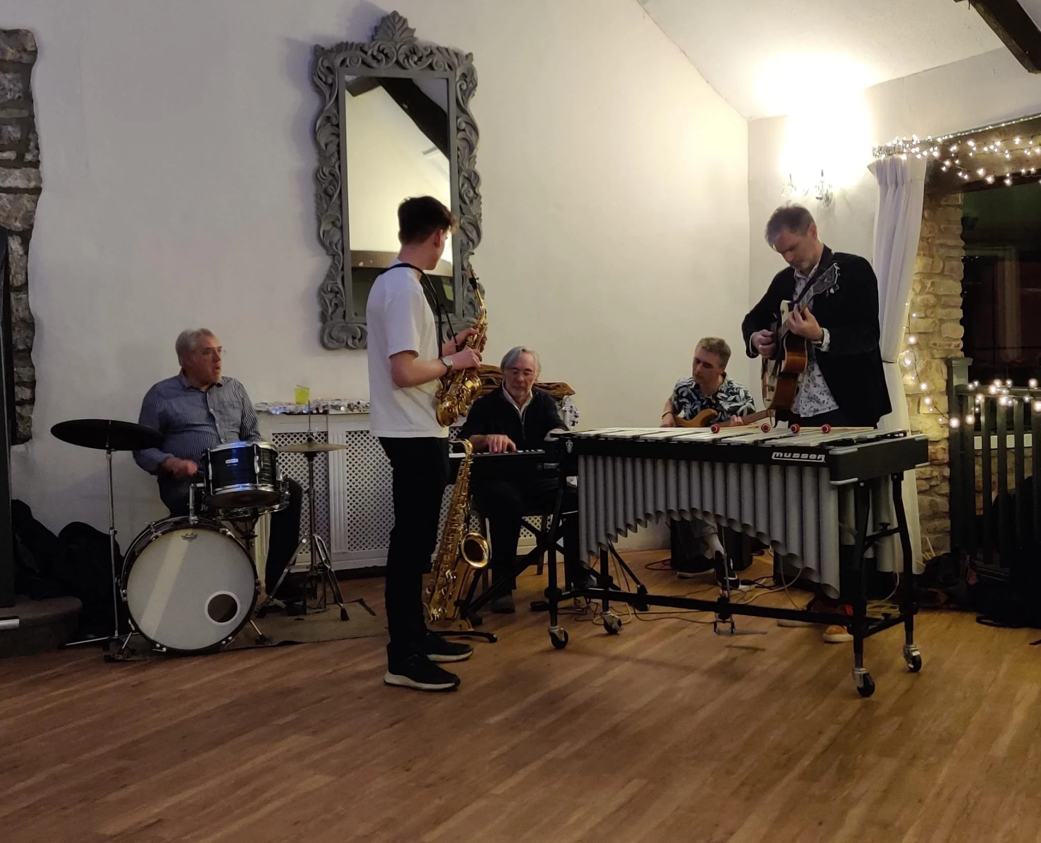 A musical band performs indoors, with five male musicians playing various instruments including drums, saxophone, keyboard, guitar, and vibraphone. The setting features a large mirror, decorative lights, and wooden flooring, suggesting an intimate or festive gathering.