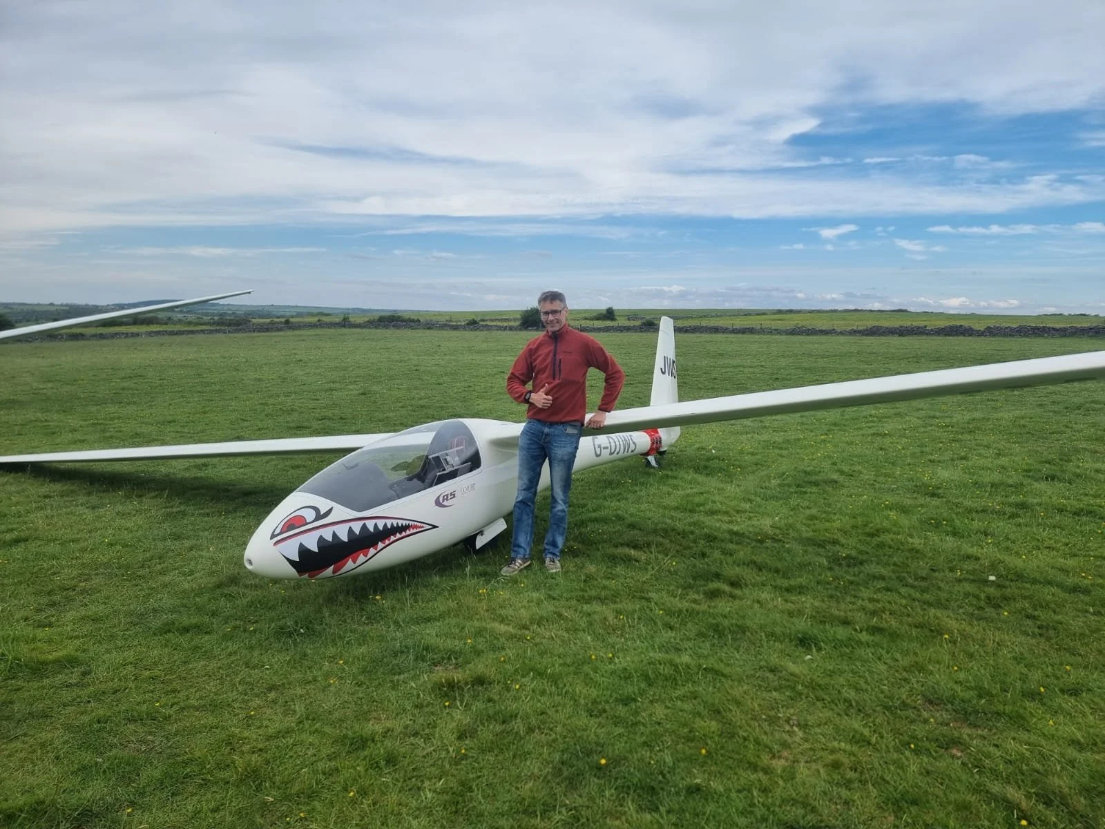 Man in red jacket standing next to a white glider plane with shark face design on the nose, on a grassy field with a partly cloudy sky in the background.