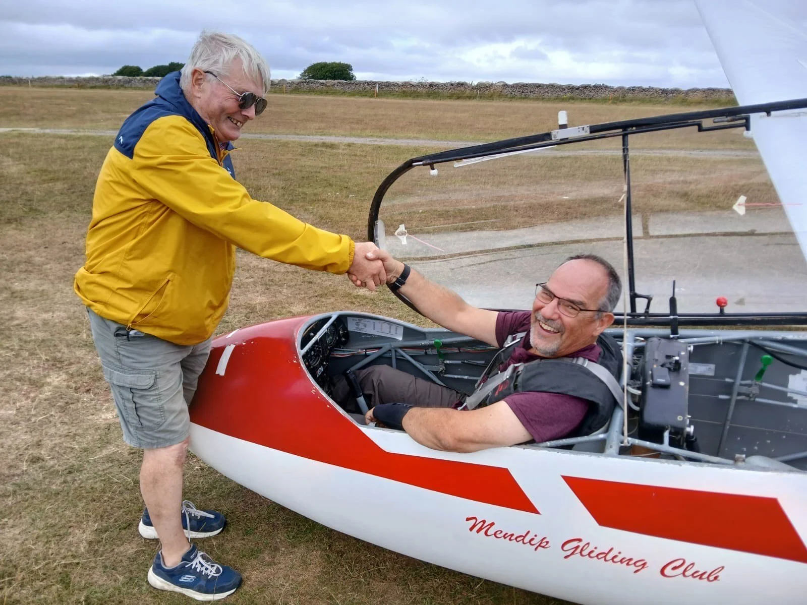 Man standing next to a small aircraft holding a red helmet, with a hangar and another small airplane in the background at an airfield.