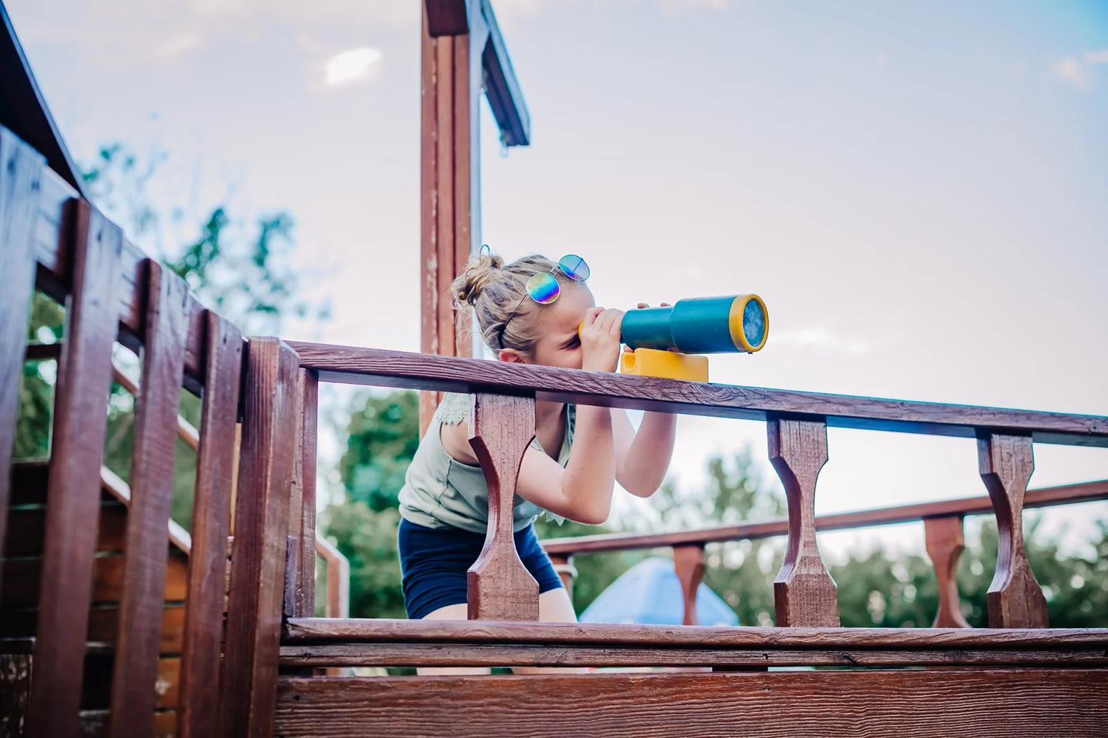 A young girl with sunglasses is looking through a telescope on a wooden deck outdoors, with trees and sky in the background.
