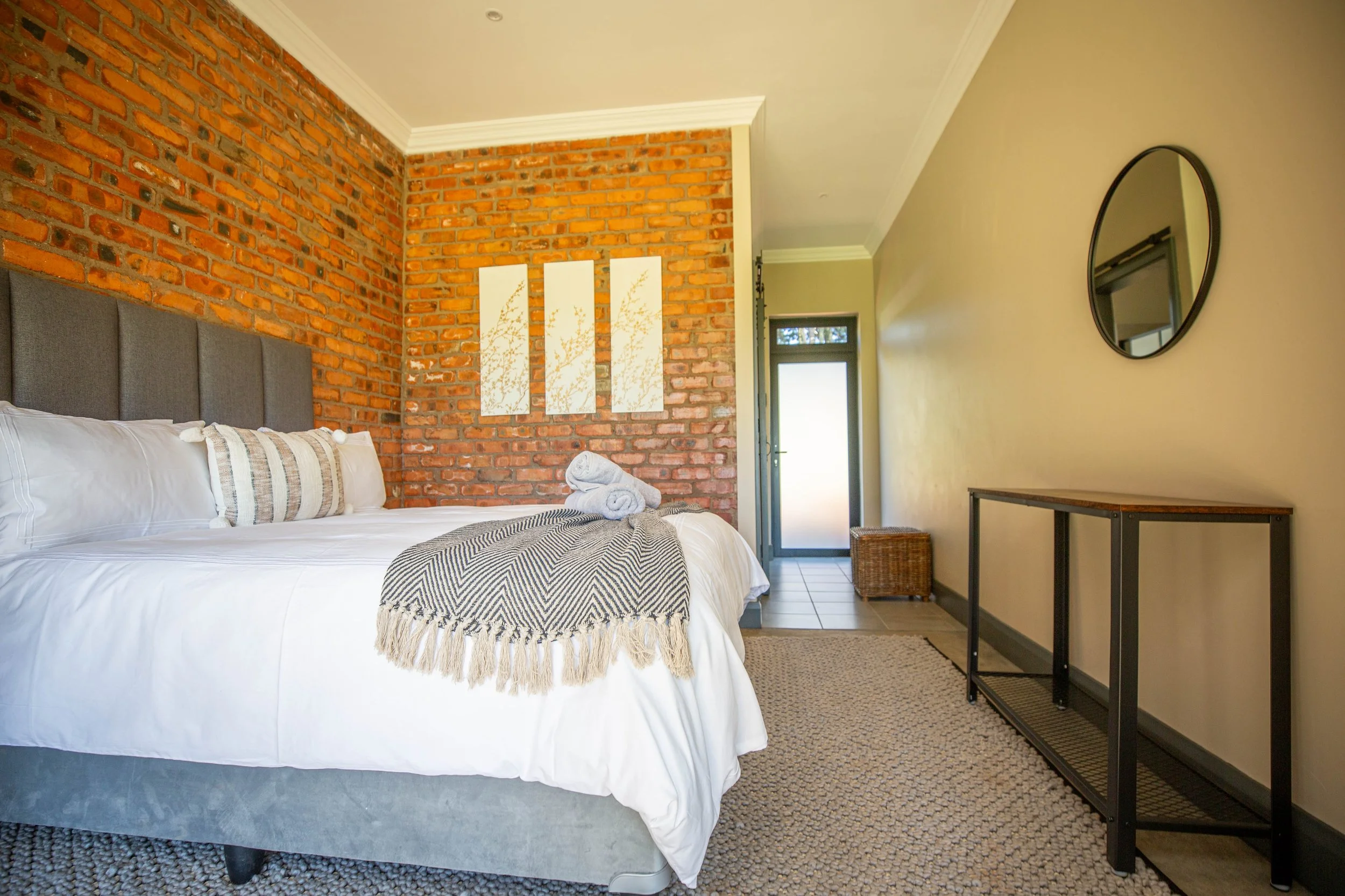 A cozy bedroom with a bed, white linens, rolled towels, a decorative pillow, a textured throw blanket, brick wall accent, modern art, a mirror, a console table, and a door leading outside.