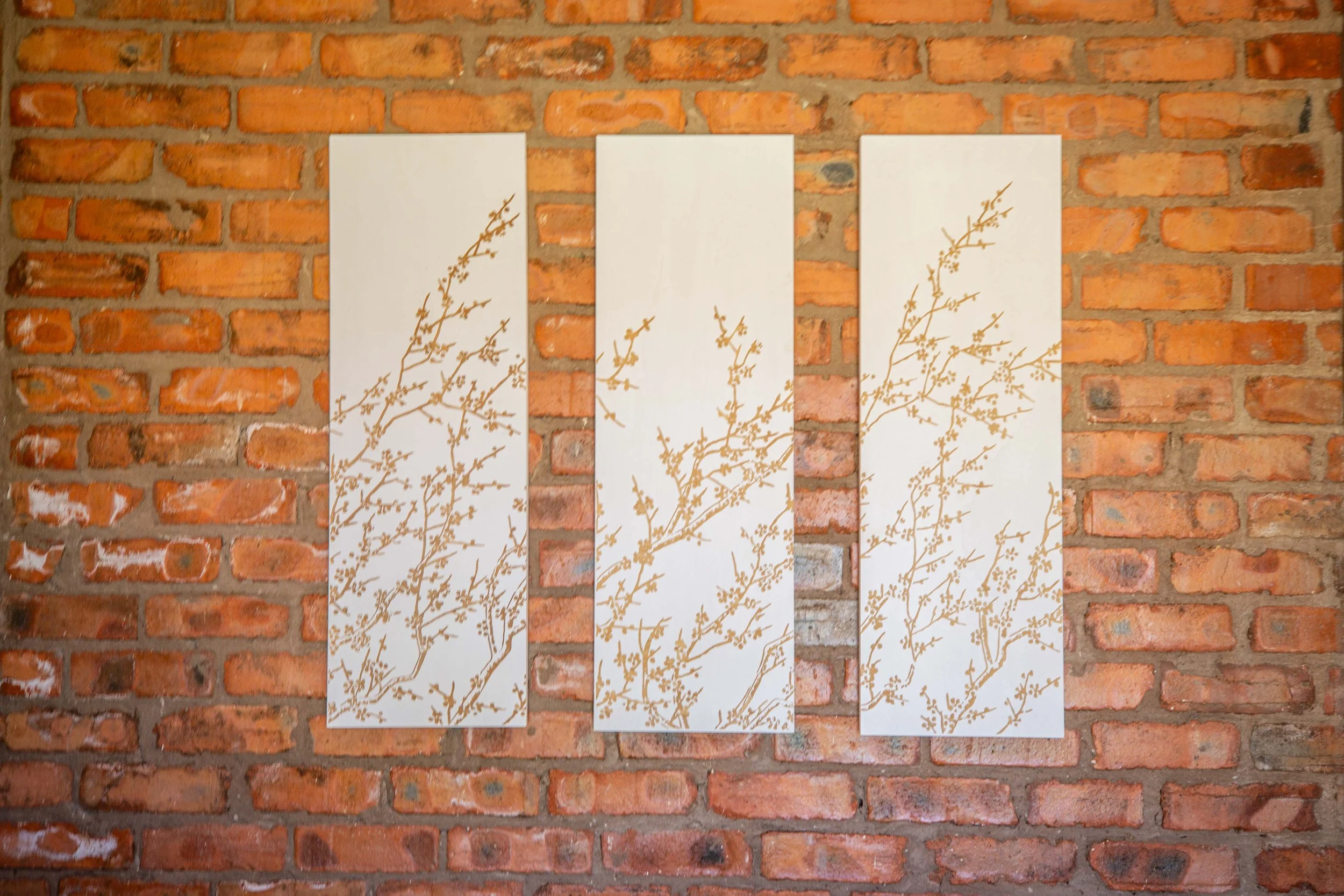 Three vertical white panels with gold branch and leaf designs hanging on a red brick wall.