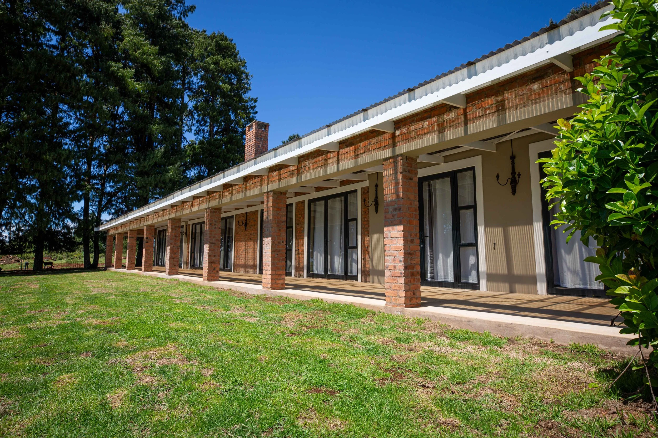 A long brick building with a covered porch, large glass doors, and a grassy yard in front, surrounded by trees.