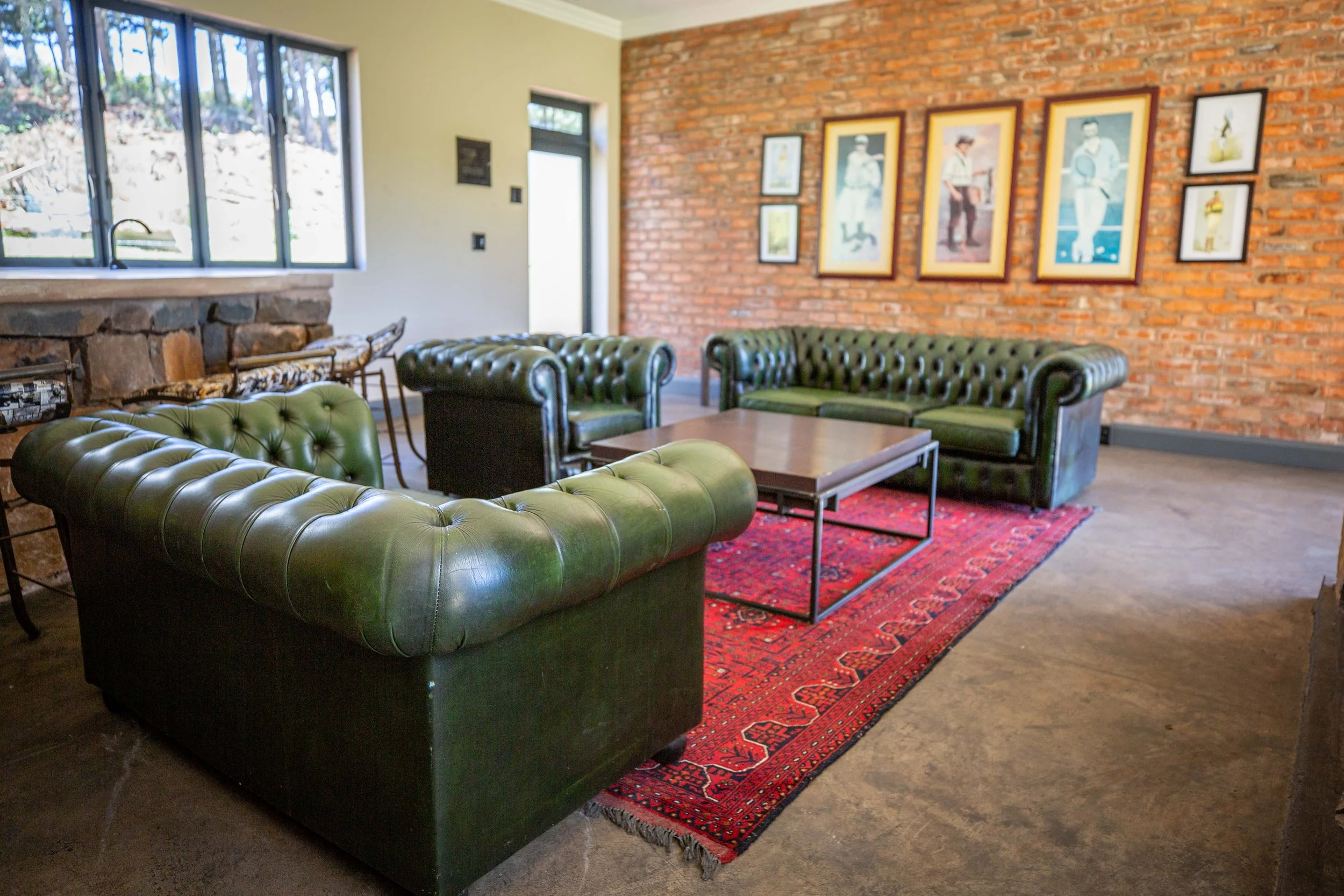 Living room with green leather Chesterfield sofas, a red patterned rug, framed artwork on a brick wall, a window, and a door.