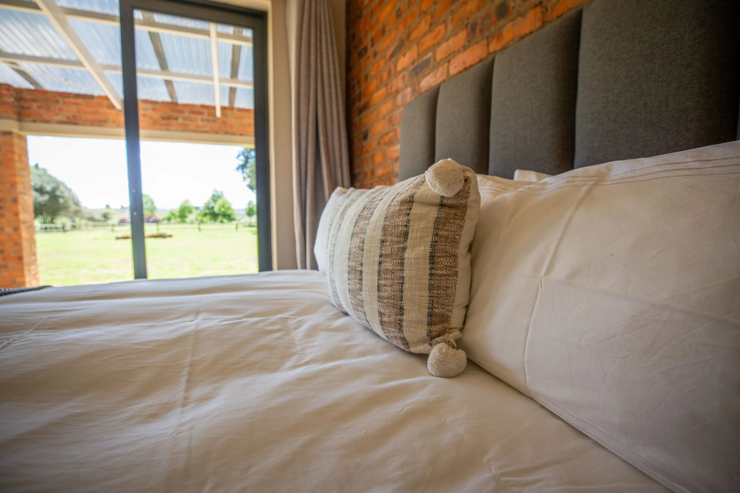Close-up of a bed with white bedding and a decorated pillow in front of a brick wall, with large glass sliding door showing an outdoor view of trees and grass.