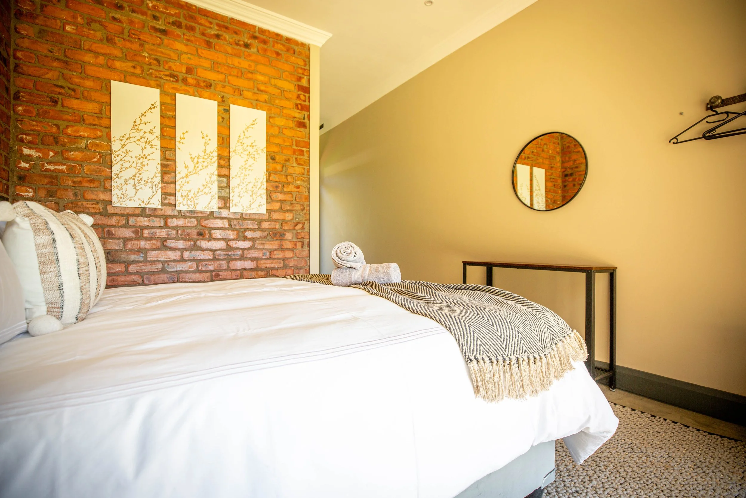A cozy bedroom with a white bed, stacked towels, a brick accent wall, a circular mirror, and wall art of white panels with gold accents.
