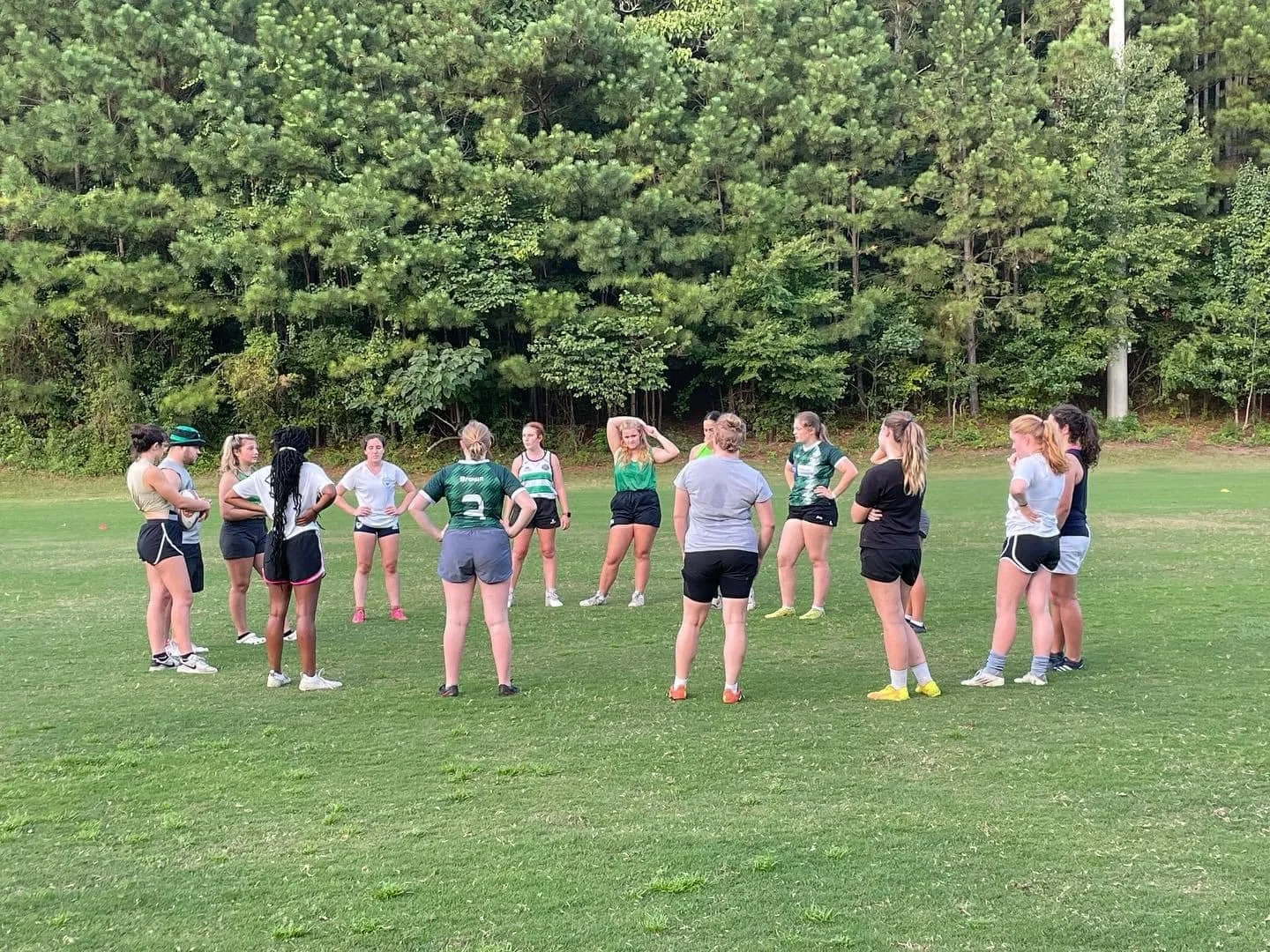 A group of young women in sportswear standing in a circle on a grassy field, with trees in the background, likely during a sports practice or team meeting.