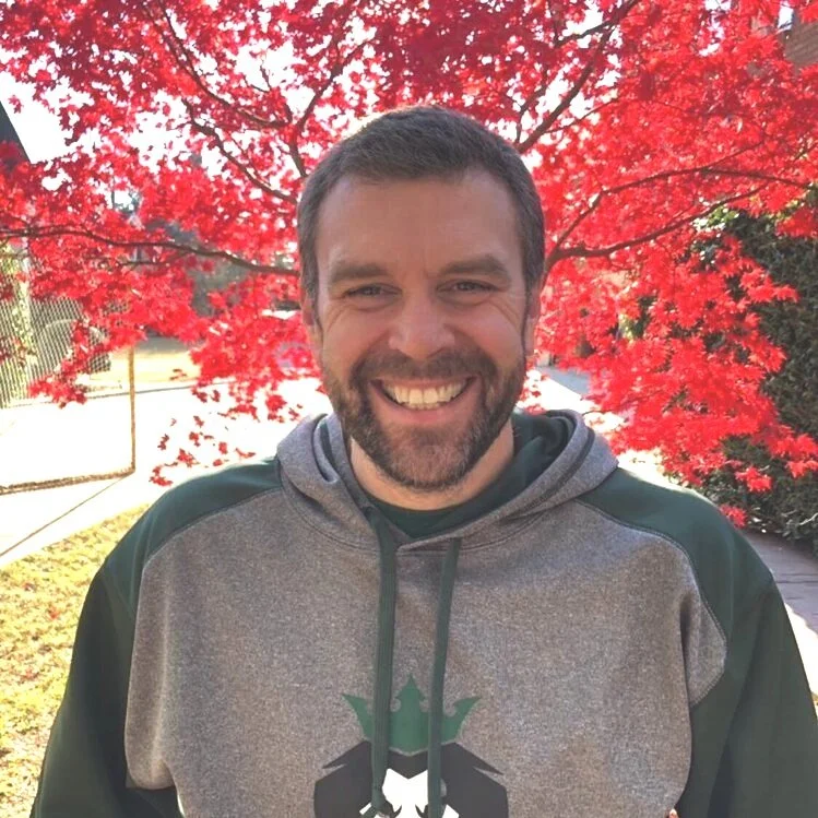 Man smiling outdoors with a vibrant red autumn tree in the background.