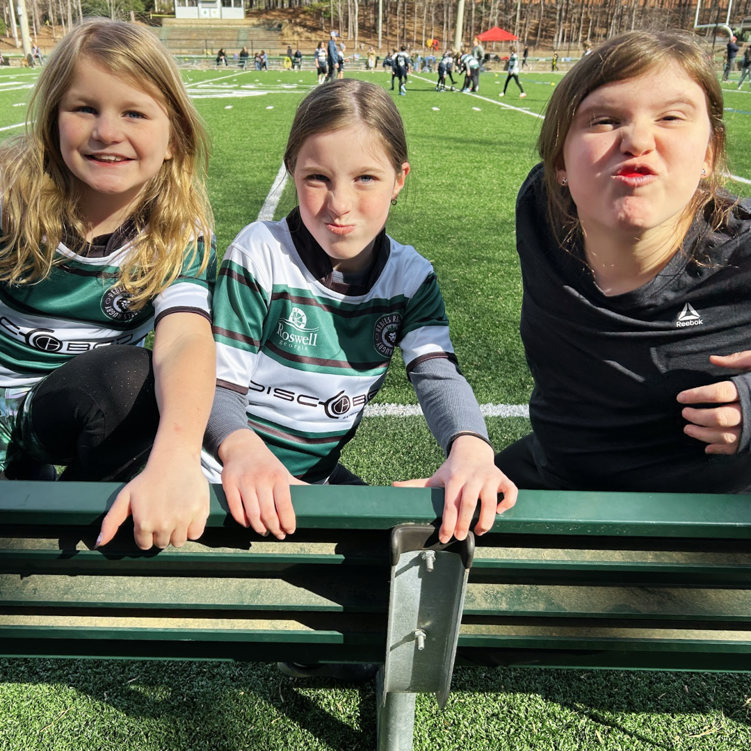Three young girls sitting on a bench on a sports field, smiling and making funny faces. Two are wearing green and white soccer jerseys, and one is wearing a black Reebok shirt. There are other people and children in the background on the field.