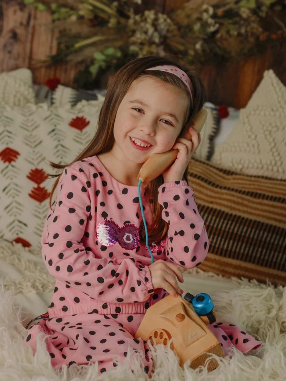 A young girl in pink pajamas with black polka dots, smiling while holding a vintage toy telephone to her ear, sitting on a fluffy white rug in a cozy room.