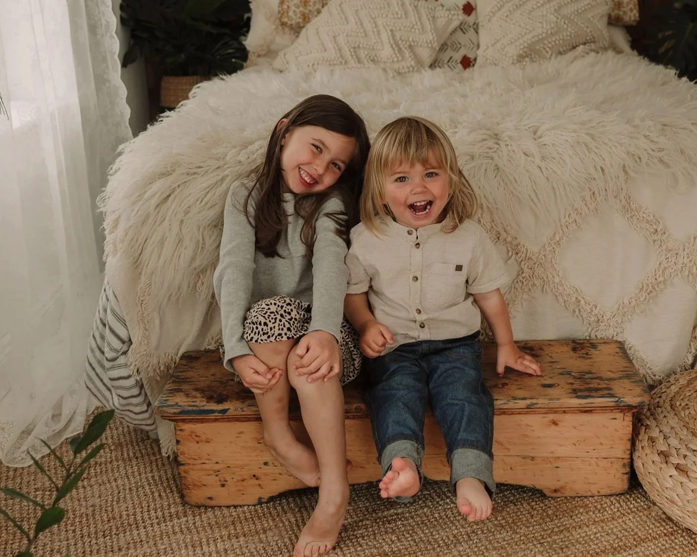 A young girl with blonde hair, wearing a white sweatshirt with red hearts and pink pants, plays with a doll in a pink patterned crib. She is standing next to a wicker doll stroller with a floral blanket. The room has wooden and pink furniture with a teddy bear in a pink box in the background, and a stuffed bunny on the floor.