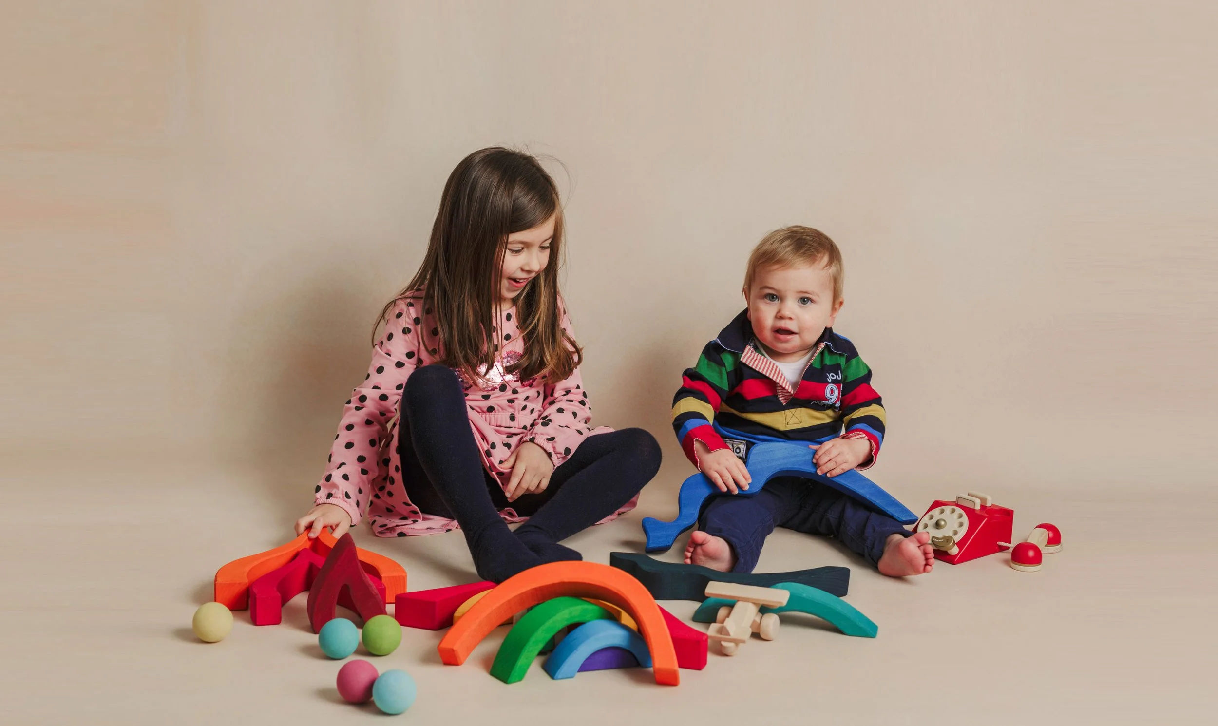 Two young children sitting on the floor playing with colorful wooden toys and balls against a plain beige background.