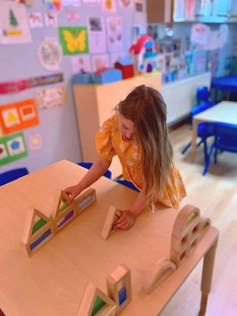 A young girl with long hair wearing an orange dress playing with wooden rainbow-colored stacking blocks on a table in a colorful classroom.
